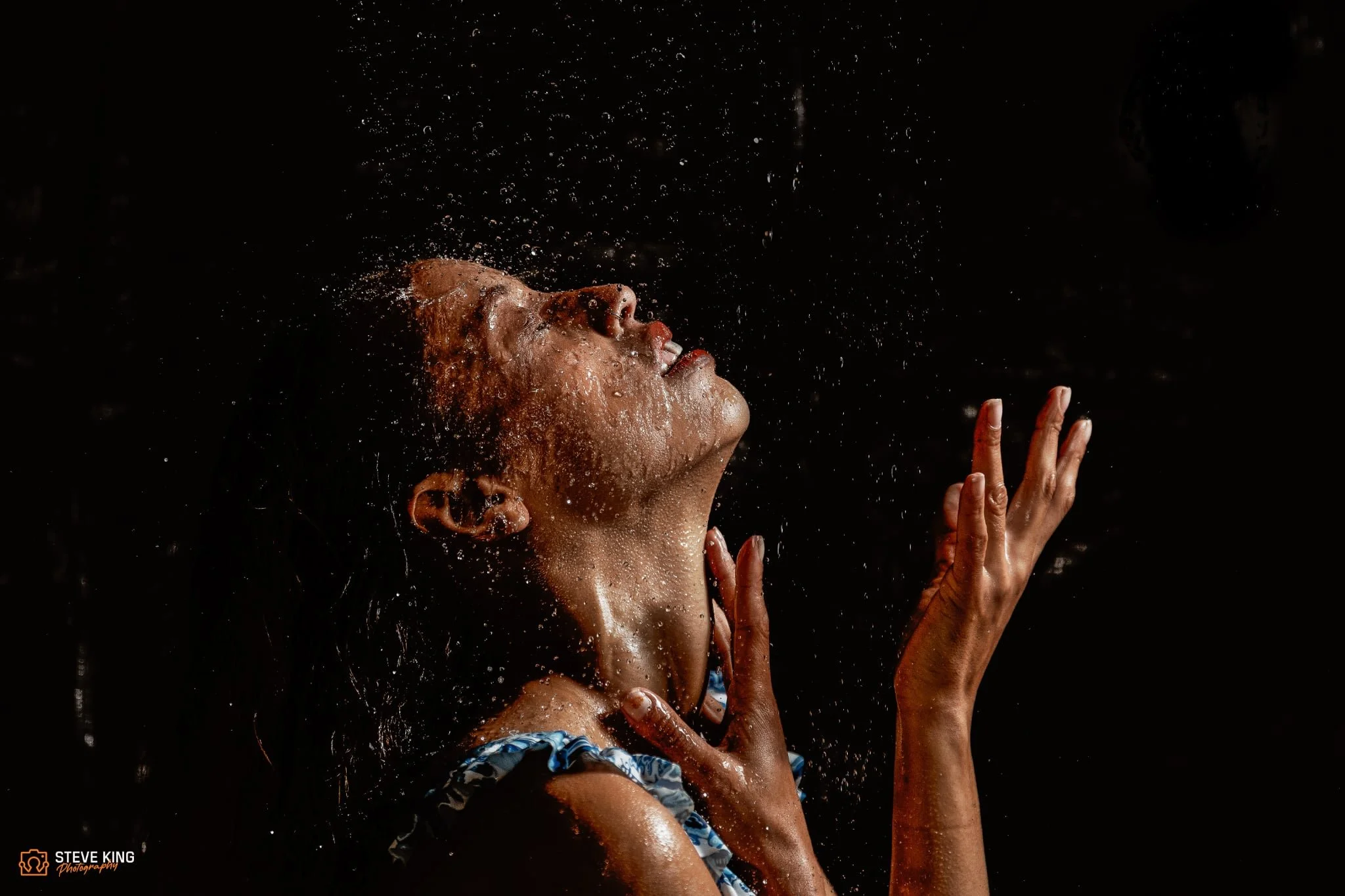 A woman with curly hair tilts her head back and raises her right hand while water splashes around her face against a black background.