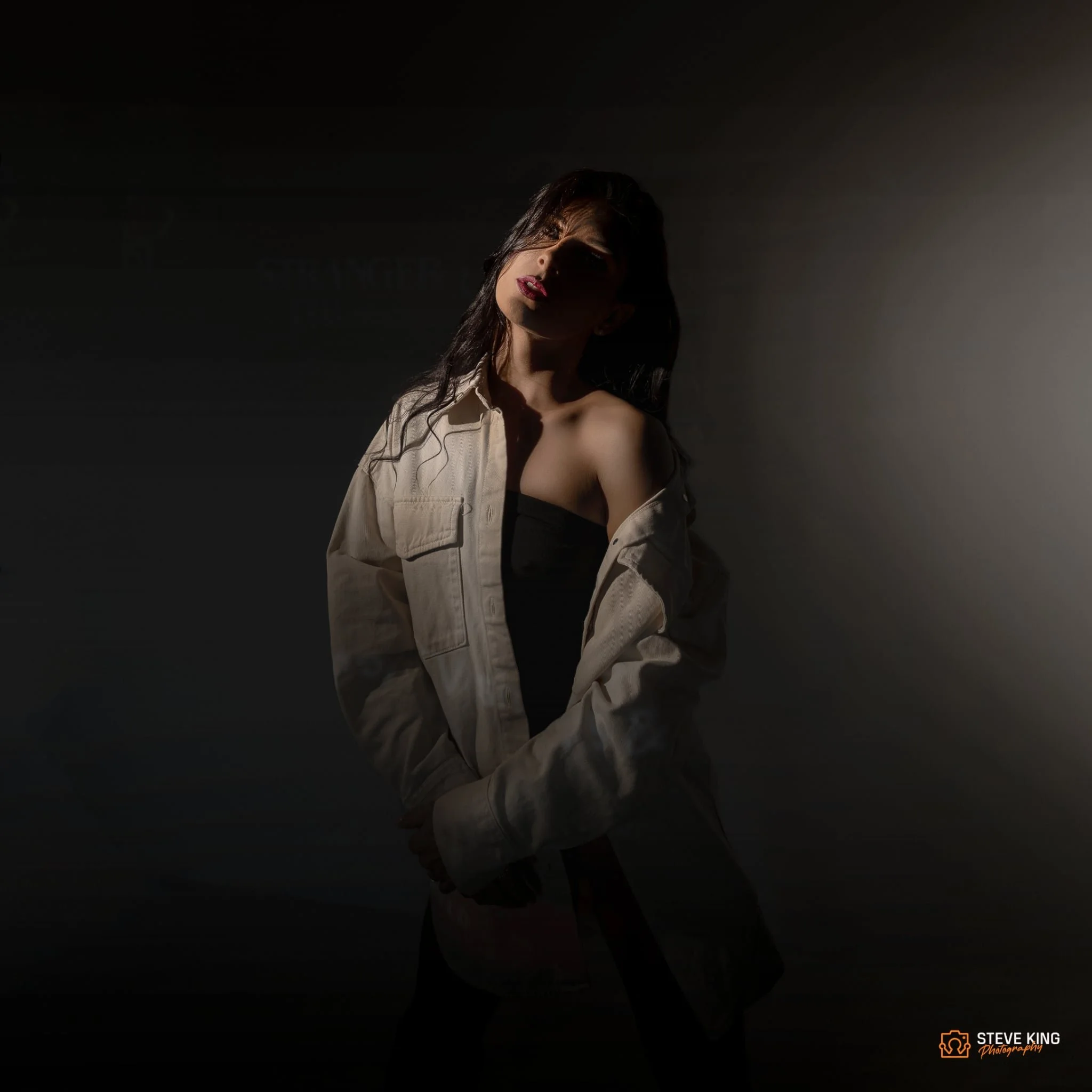 Young woman with dark hair and red lipstick, wearing a black top and beige jacket, standing against a dark background with dramatic lighting.