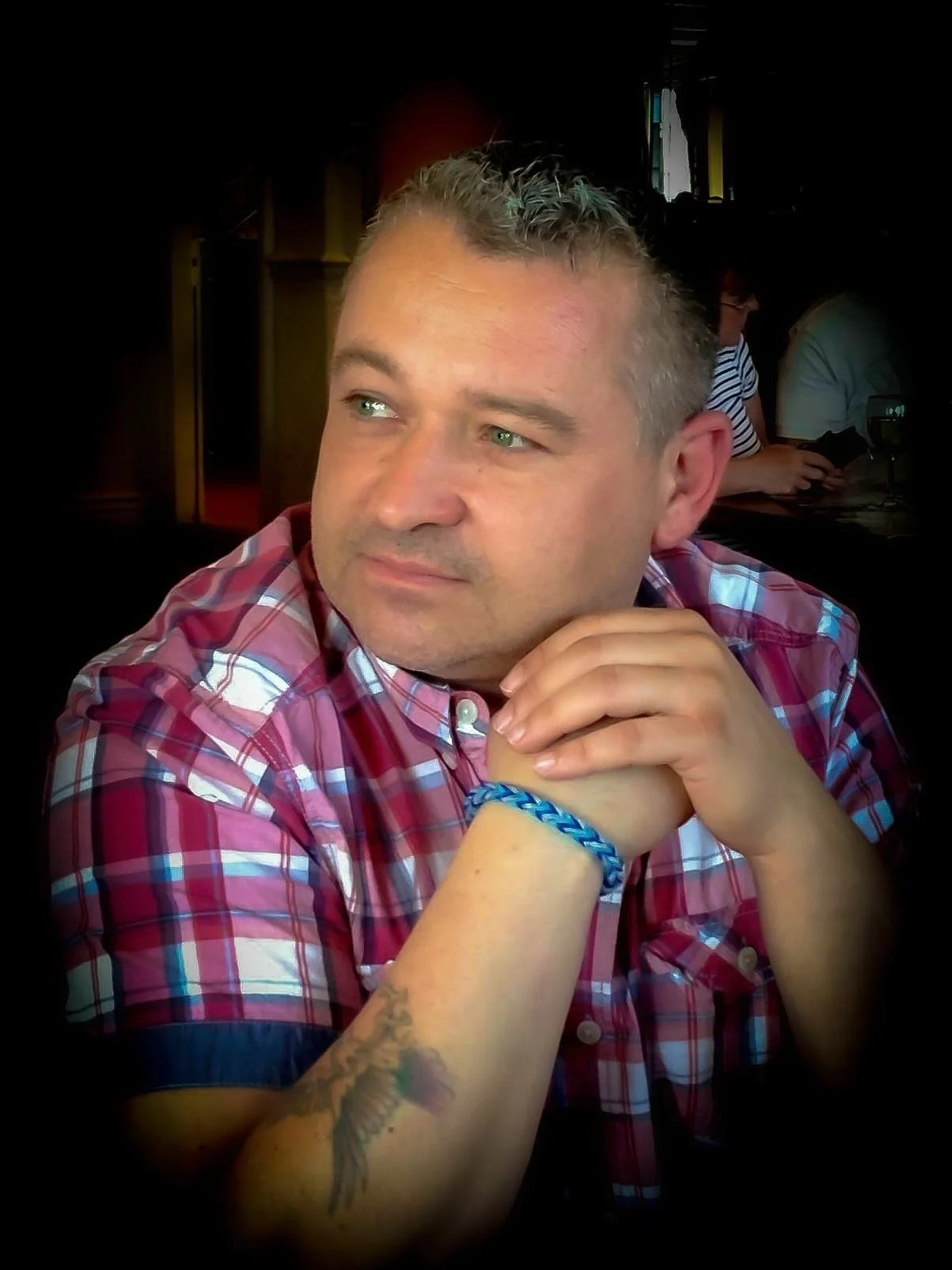 A man with short blond hair and green eyes wearing a red plaid shirt, sitting at a table in a restaurant or cafe. He is resting his chin on his hands, with a blue braided bracelet on his wrist and a tattoo of a bird on his forearm. In the background, there are other people and a window with natural light.