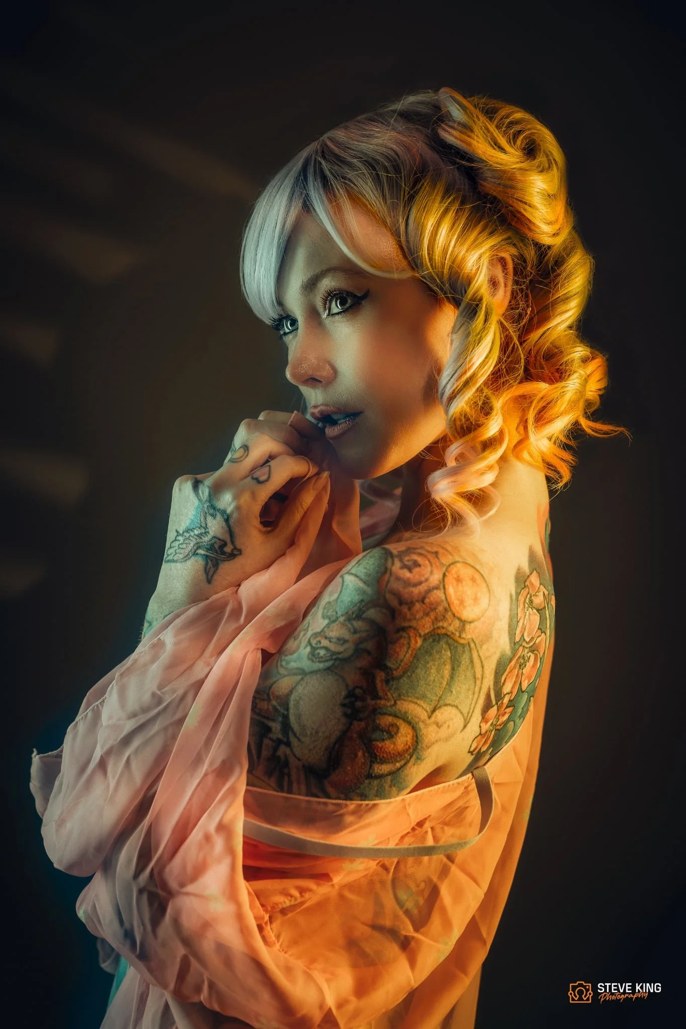 Portrait of a woman with colorful tattoos on her arm and shoulder, wearing a sheer orange fabric, with platinum blonde hair styled in large curls, looking thoughtfully to the side with hand near her face, against a dark background.