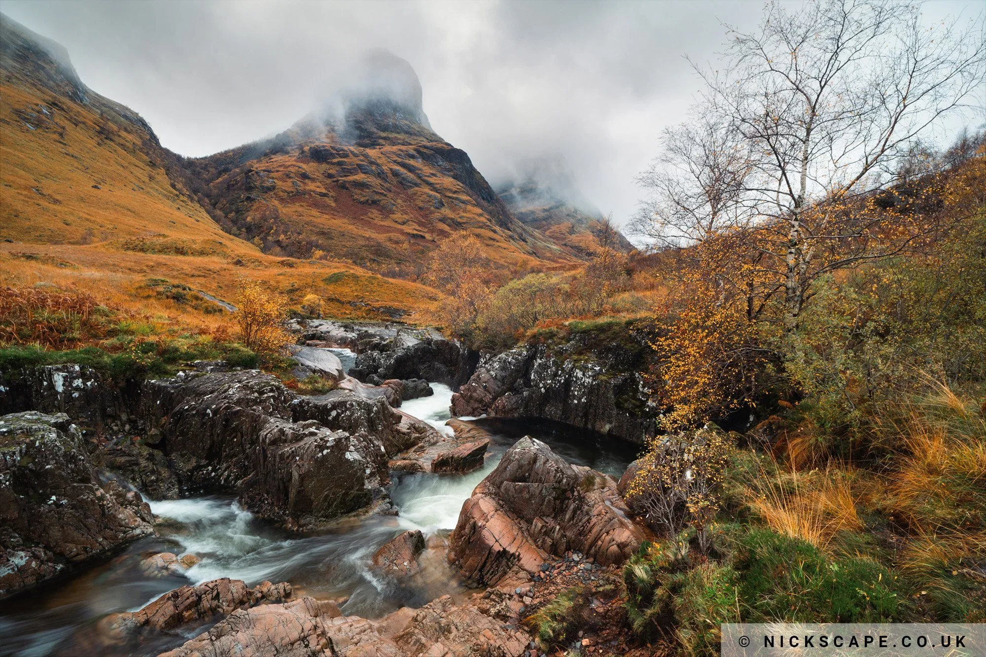 A scenic view of a mountain landscape with a river flowing over rocks, surrounded by autumn-colored foliage and misty clouds at the mountain peaks.