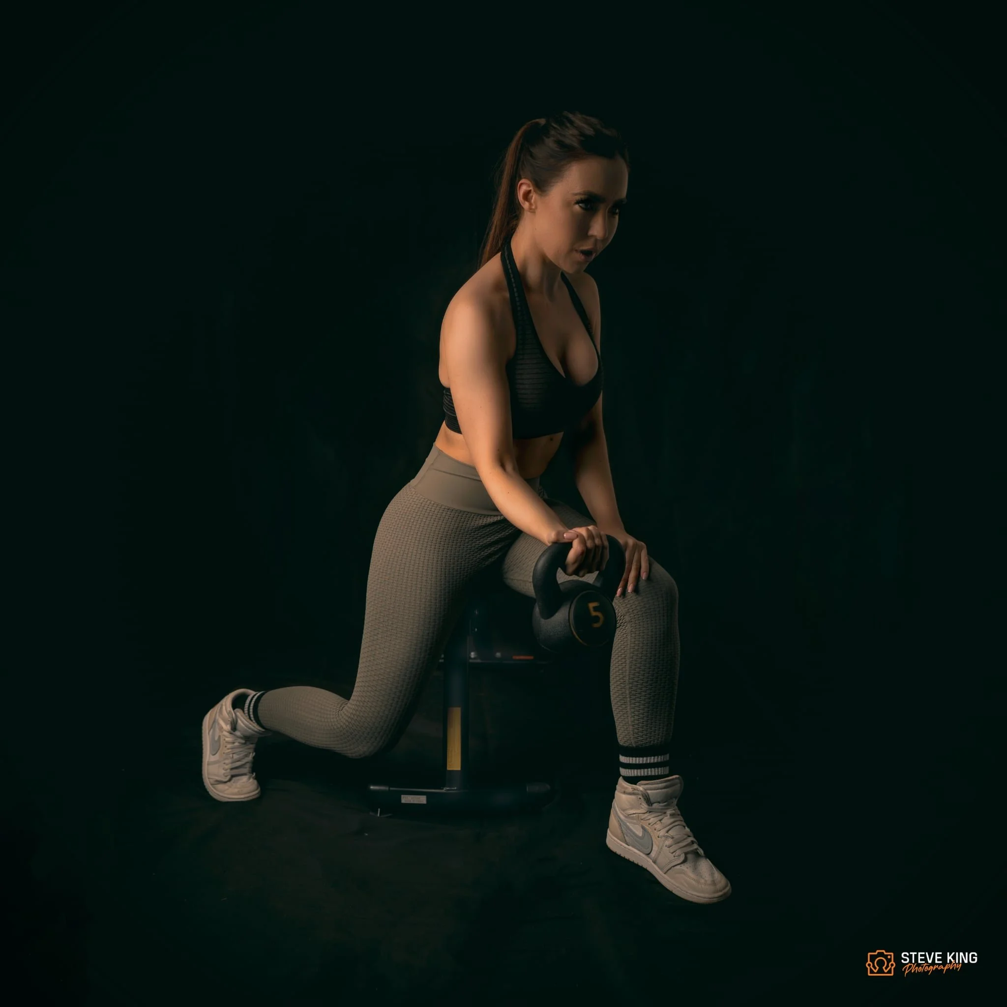A woman in athletic wear performing a lunge exercise with a kettlebell in a dark studio setting.