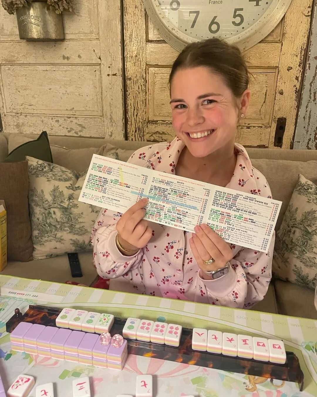 A woman sitting at a table holding up a mahjong card and smiling. There are Mahjong tiles and dice on the table in front of her. She is in a room with rustic decor, including a clock on the wall.