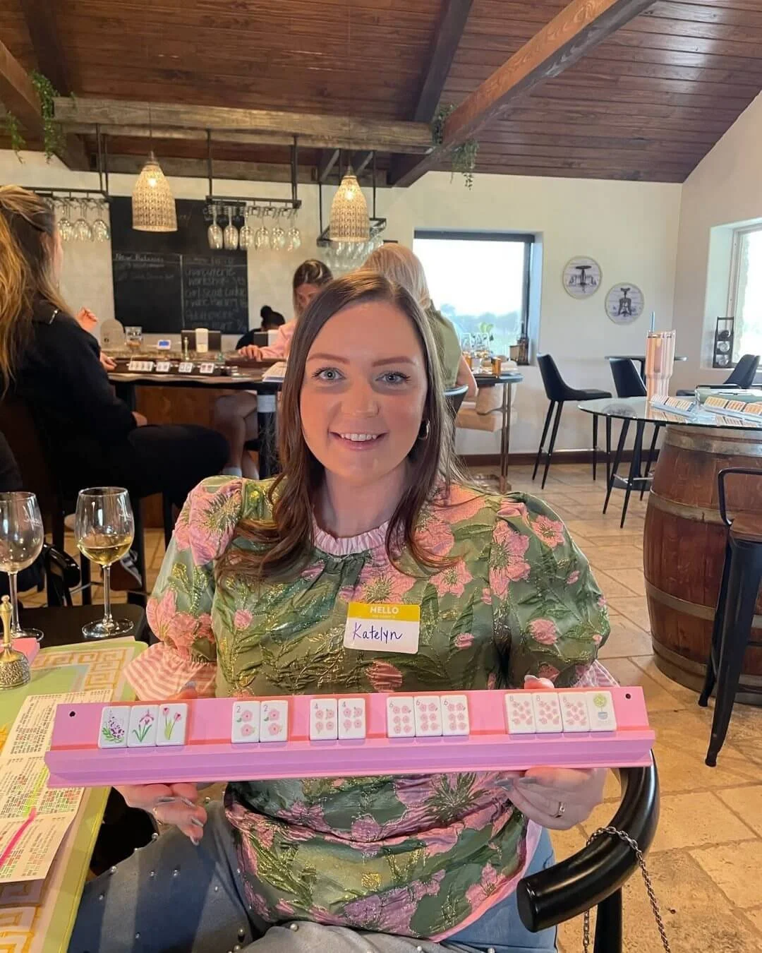 A woman with shoulder-length brown hair and a floral blouse holding a pink Mahjong tile rack at a restaurant or cafe. The background shows other patrons, hanging pendant lights, and a chalkboard menu.