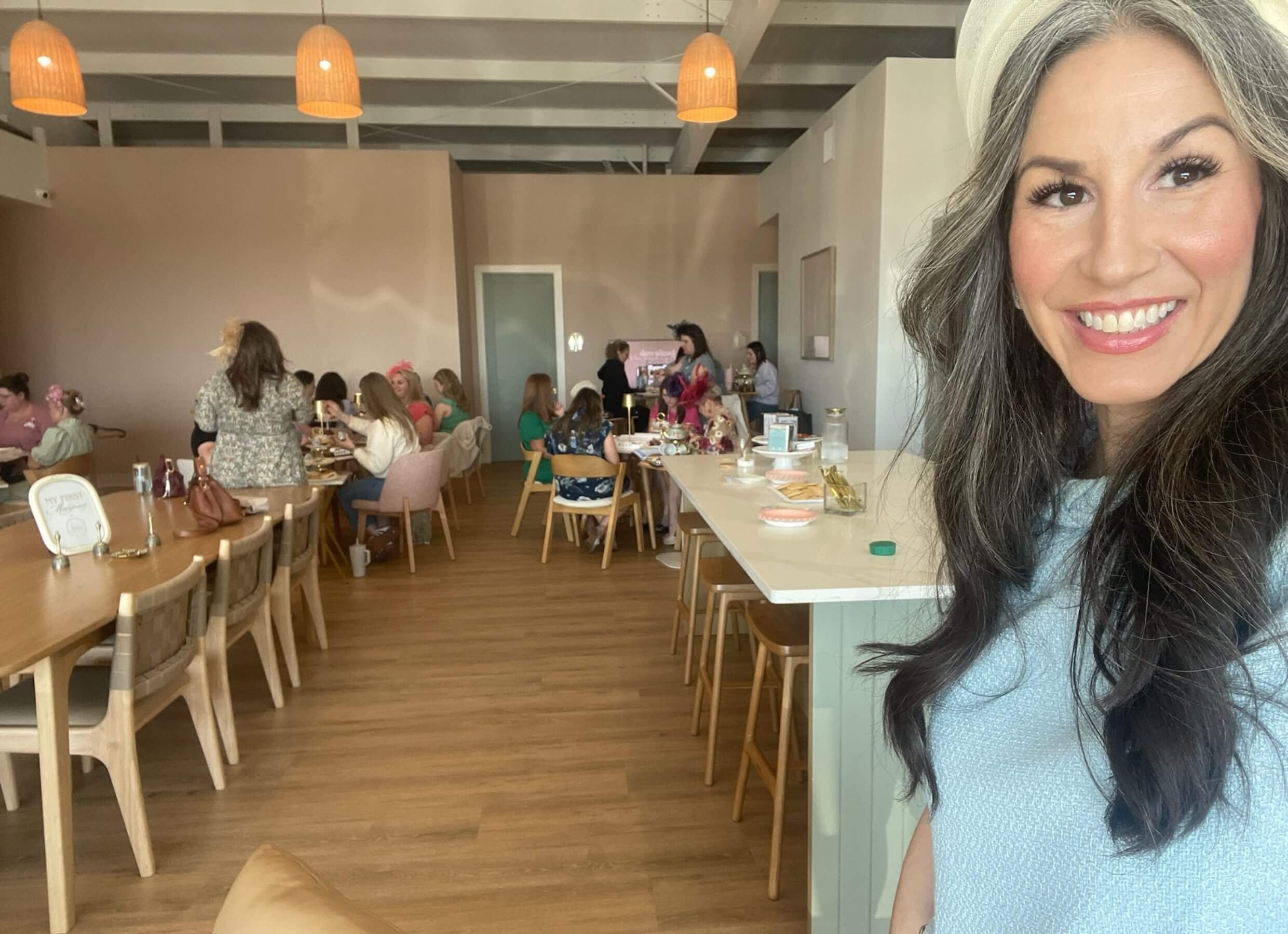 A woman with long dark hair, smiling, wearing a light blue top, standing in a busy cafe or restaurant with people sitting at mahjong tables and a beige wall in the background.