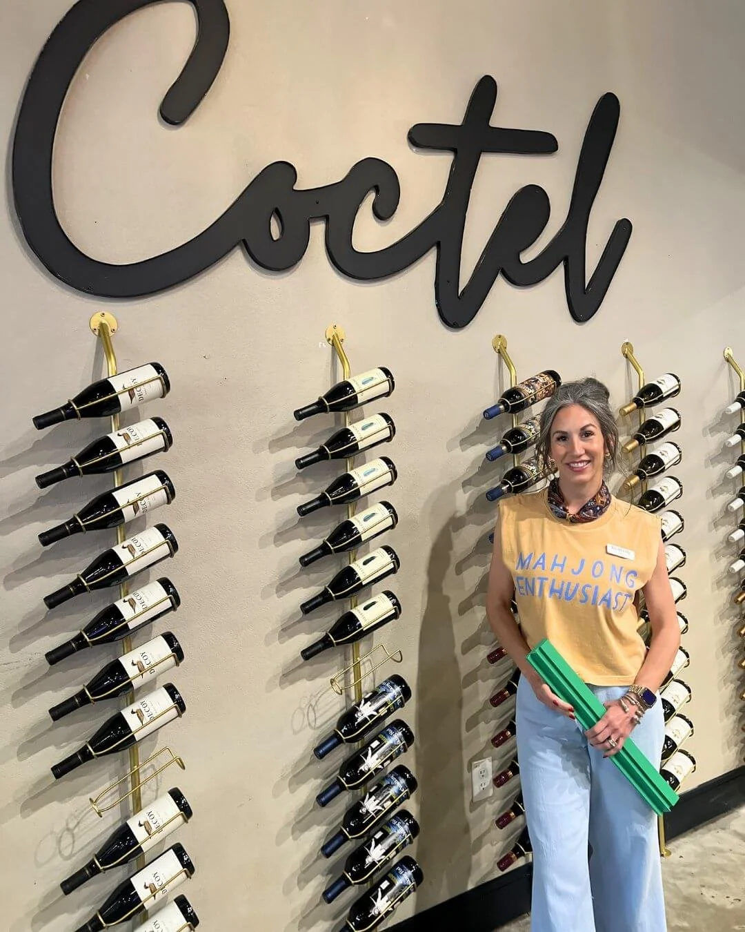 A woman standing in front of a wall with wine bottles on wine racks, with a large cursive sign above that says 'Coctel'.