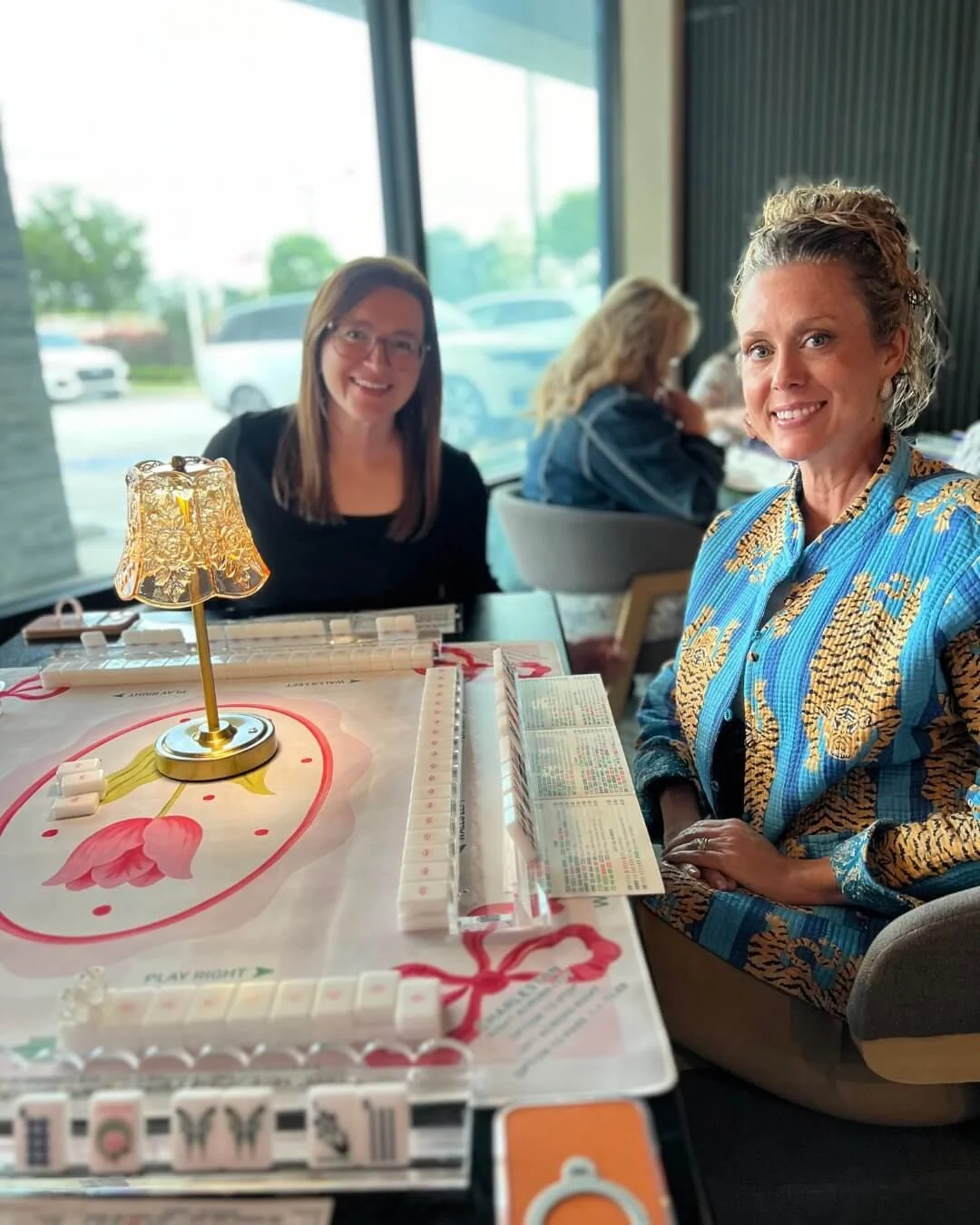 Two women sitting at a table playing a game of Mahjong in a restaurant or cafe with large windows, other patrons, and parked cars outside.