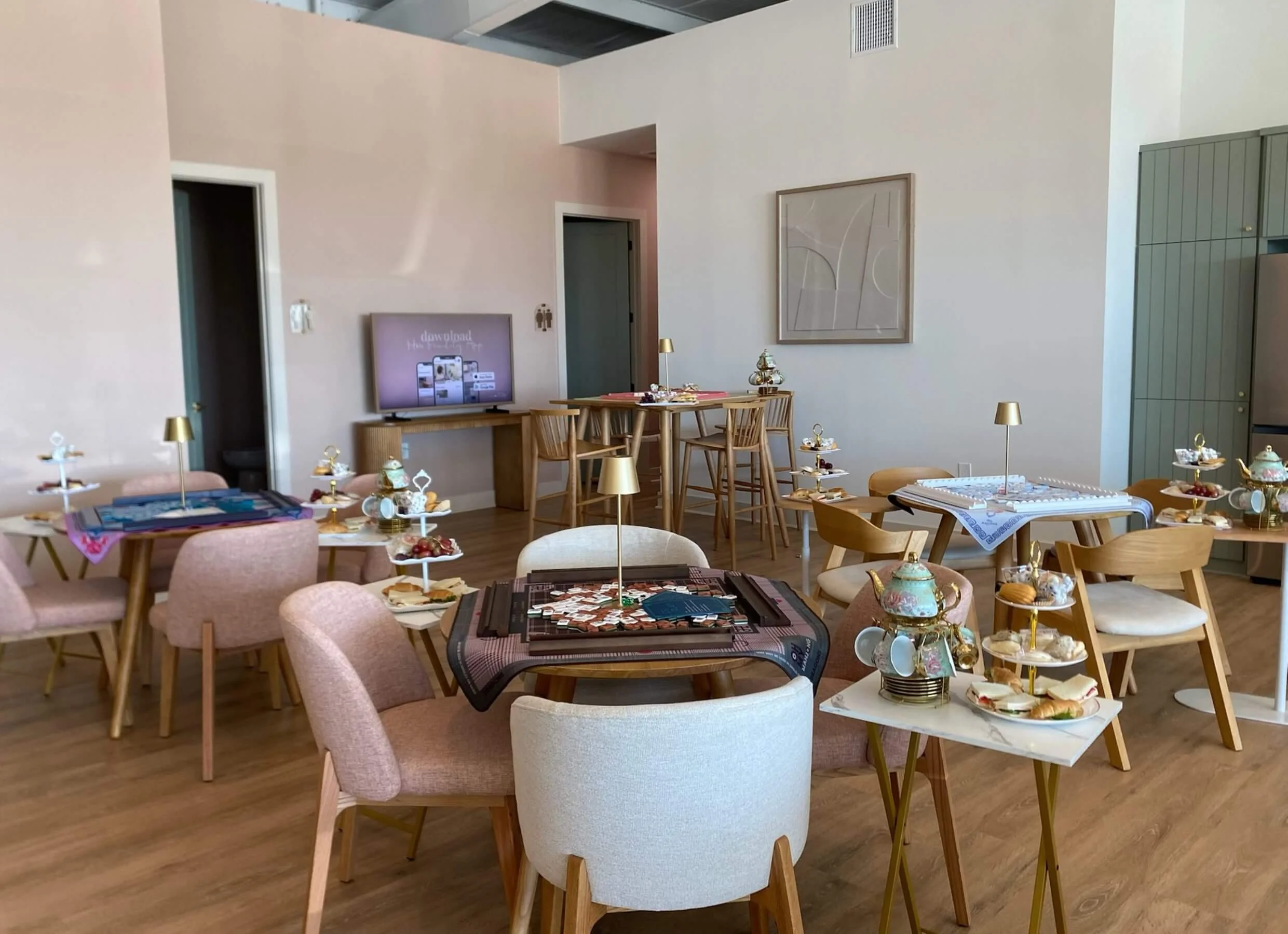 A room set up for a tea party with pastel-colored chairs, tables with desserts, and decorative mahjong boards and tiles. There is a flat-screen TV on the wall and artwork hanging nearby. The space has wooden flooring and modern decor.