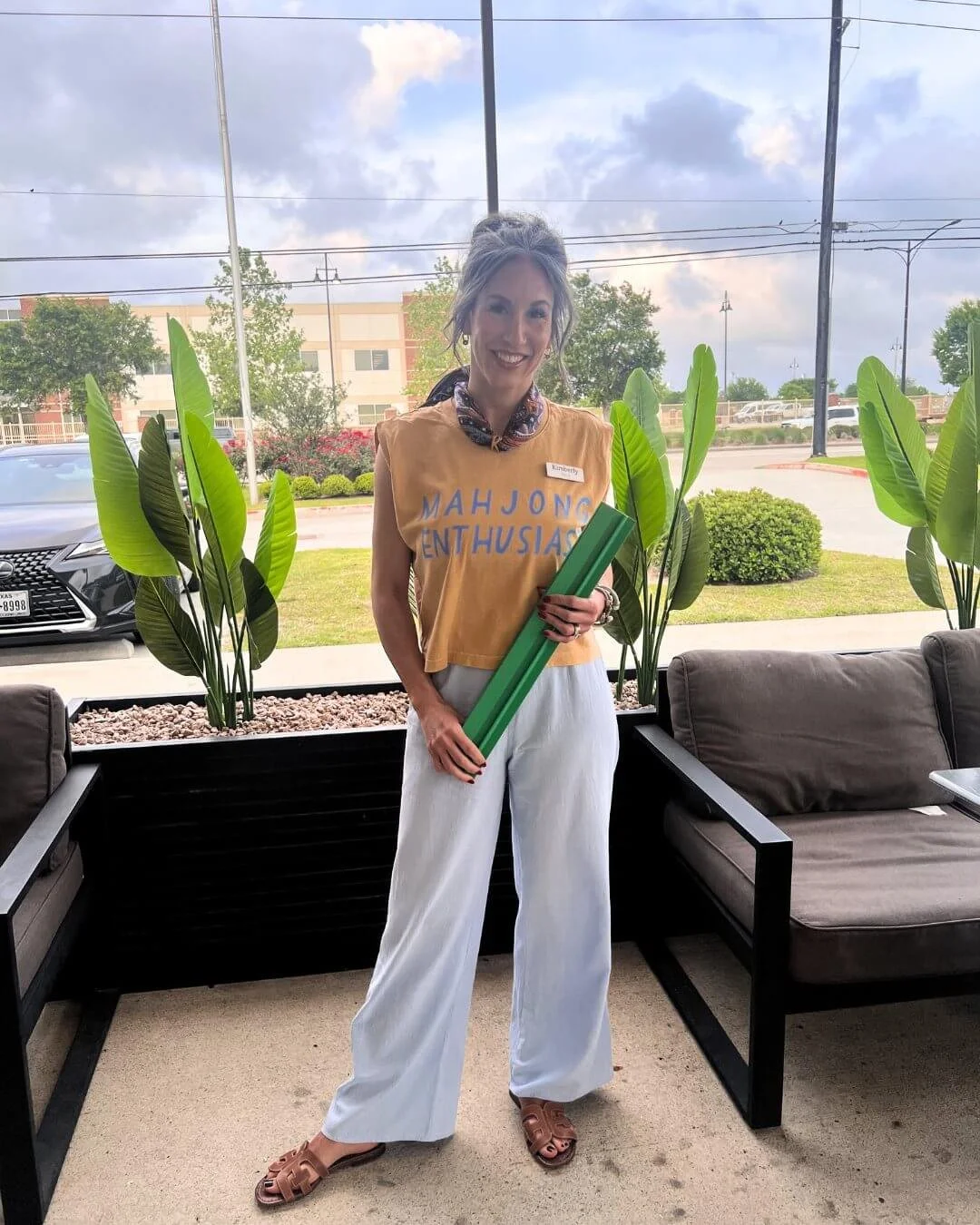 A woman with a name tag reading Kimberly, standing inside in front of large windows, holding a green foam baton, with potted plants and a parking lot outside.
