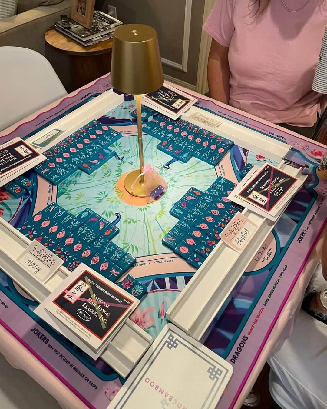 A family playing Mahjong around a table with a pink and blue patterned tablecloth. The Mahjong game has blue tiles with pink and white designs and is framed by white plastic borders. There is a small lamp with a gold shade in the center of the table, and the players' names are visible on the table surface.