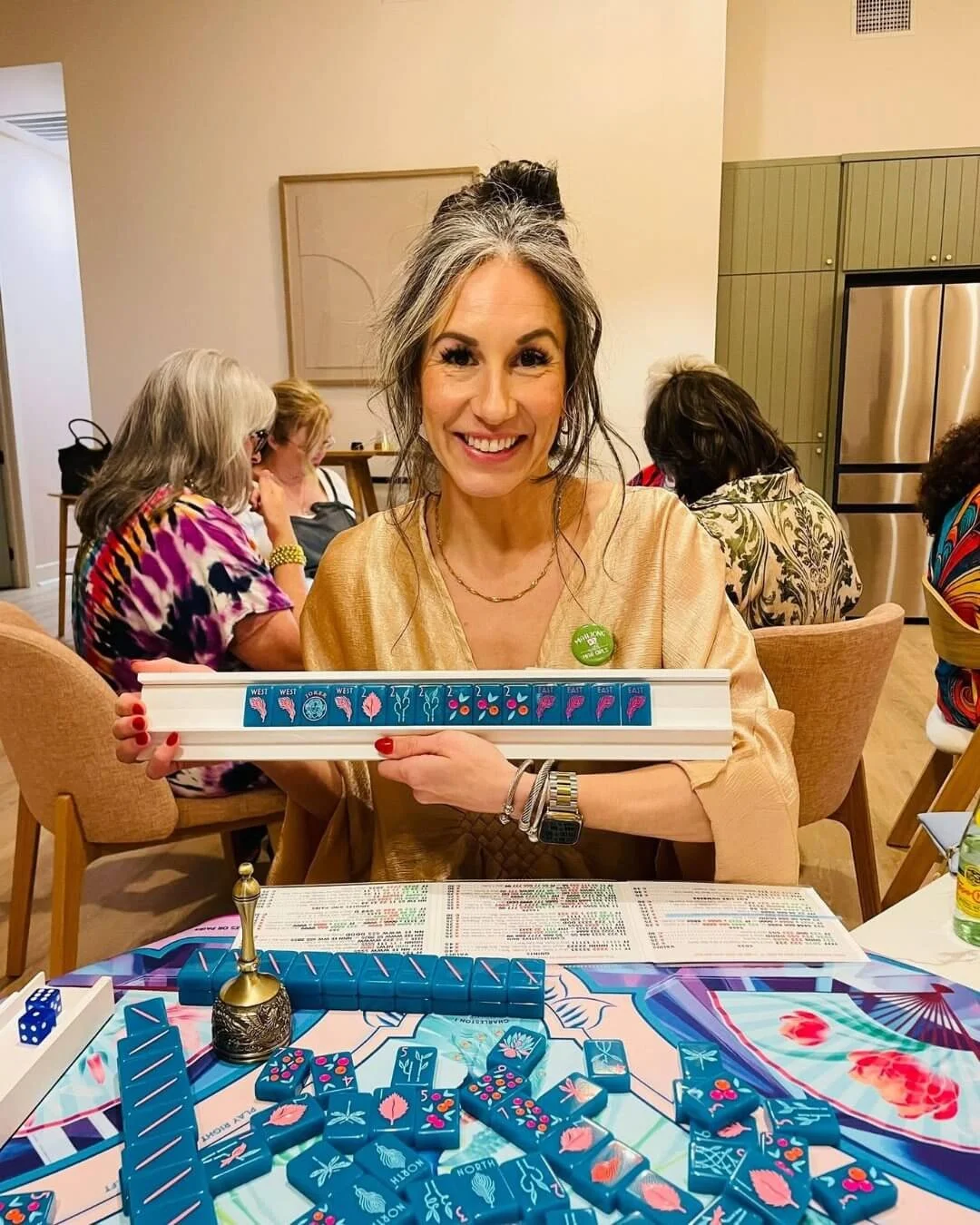 Woman smiling and holding a framed set of Mahjong tiles, sitting at a table with Mahjong game pieces, in a room with other women in the background.