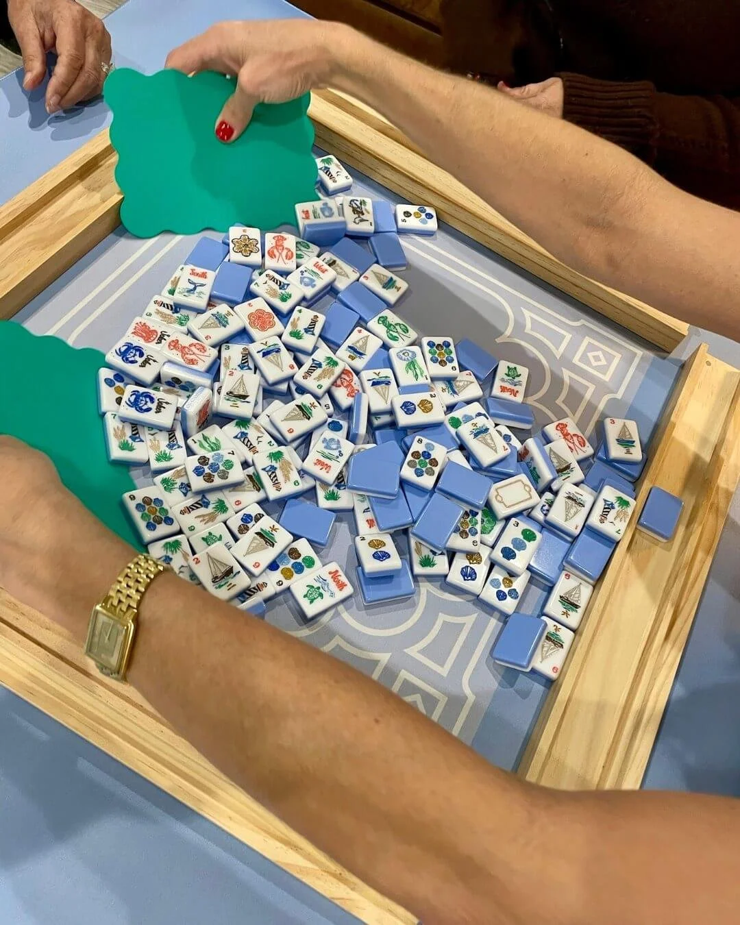 People playing Mahjong with tiles on a wooden tray. The tiles have various colorful symbols and photos.
