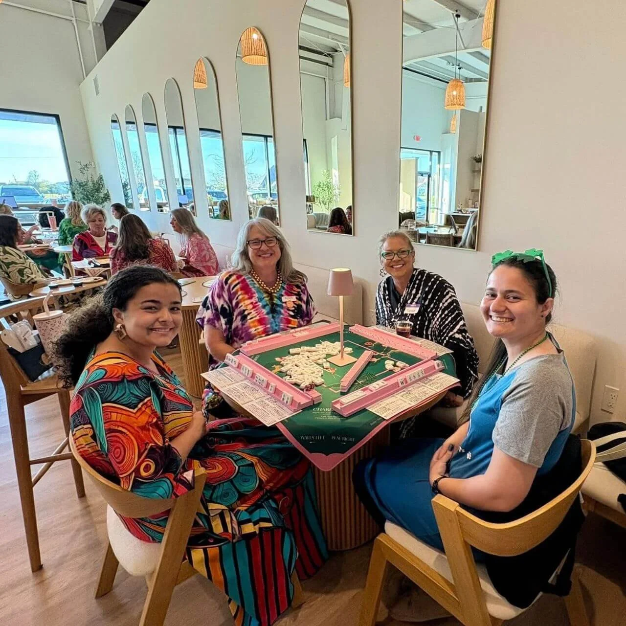 Four women sitting around a table playing a game of Mahjong in a brightly lit restaurant. They are smiling and having a good time, with other diners visible in the background.