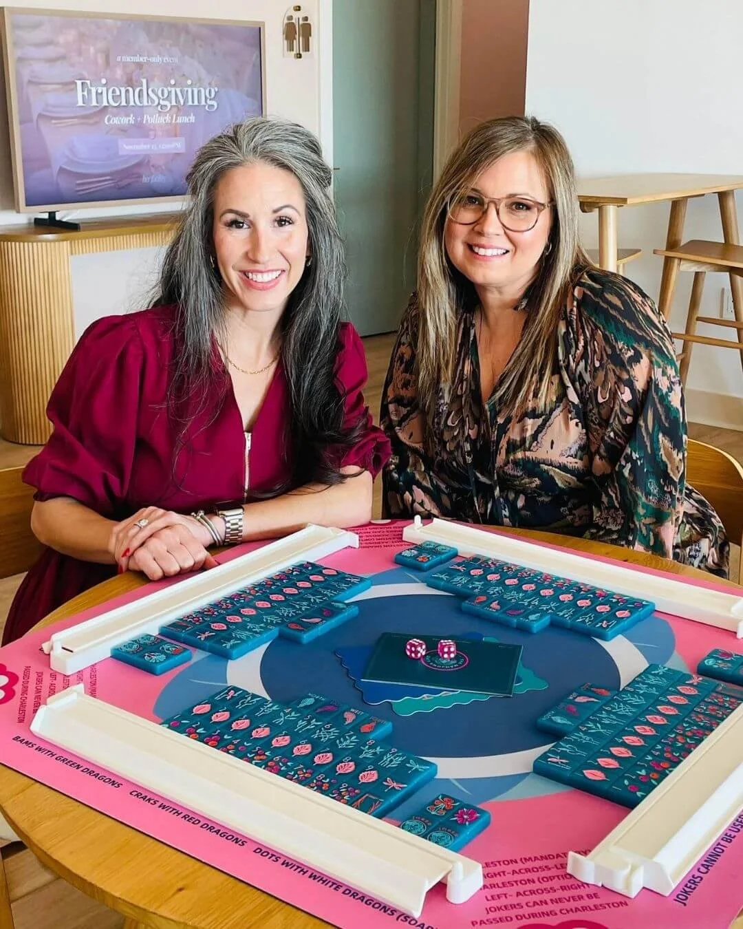 Two women playing a game on a pink and blue table, with dominoes and dice, in a room with a television in the background displaying 'Friendsgiving Lunch'.