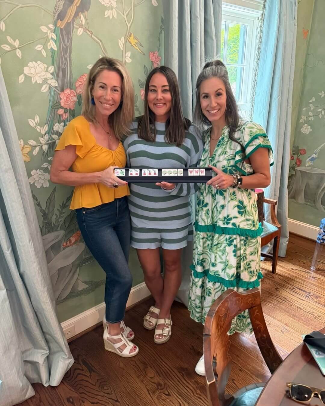 Three women smiling and holding a Mahjong tile tray in a brightly decorated room.