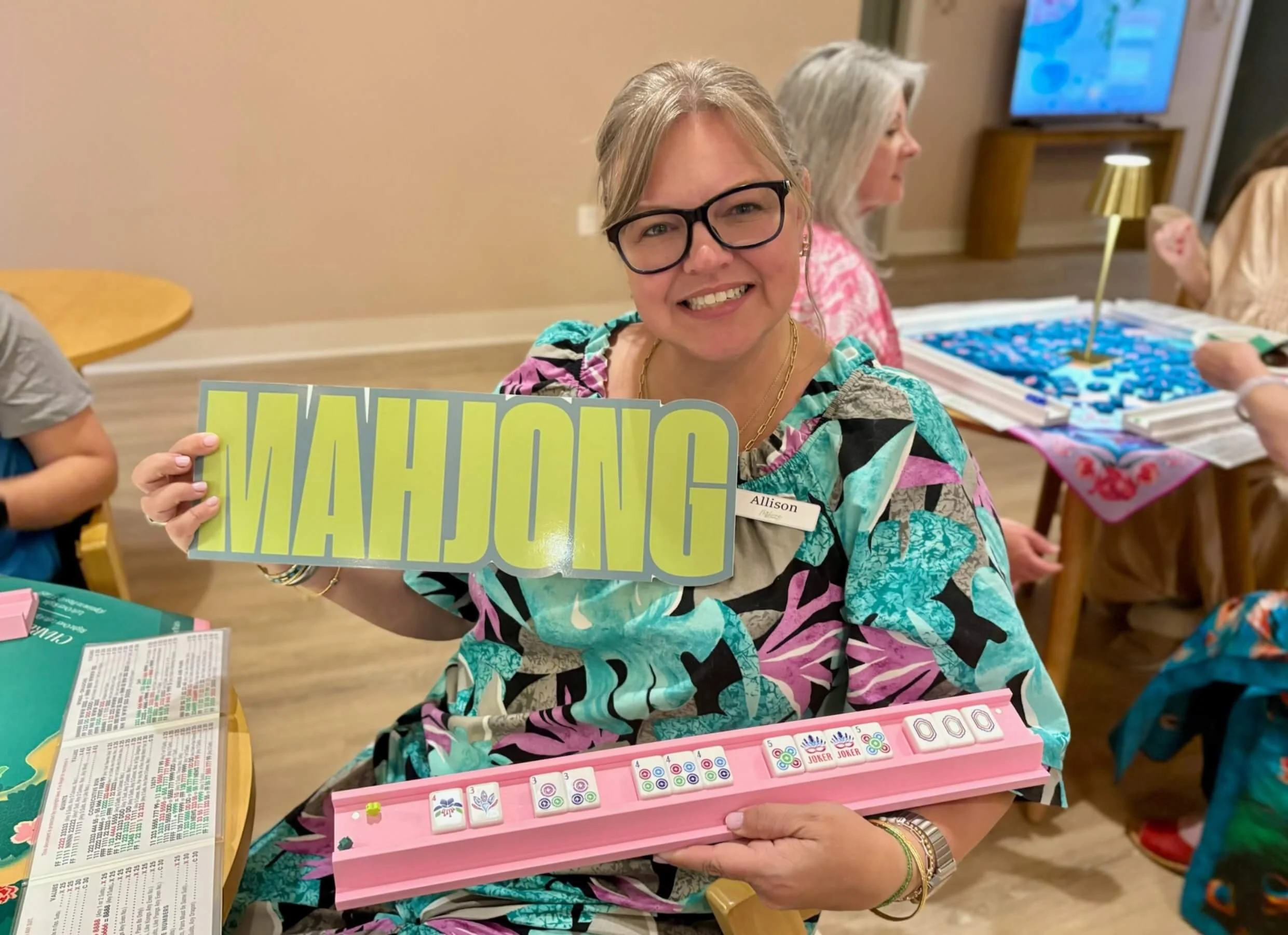 A smiling woman with glasses and a colorful patterned shirt holds a sign that says 'MAHJONG' in a lively indoor setting. She is seated at a table with various games and papers, with other women and children in the background.
