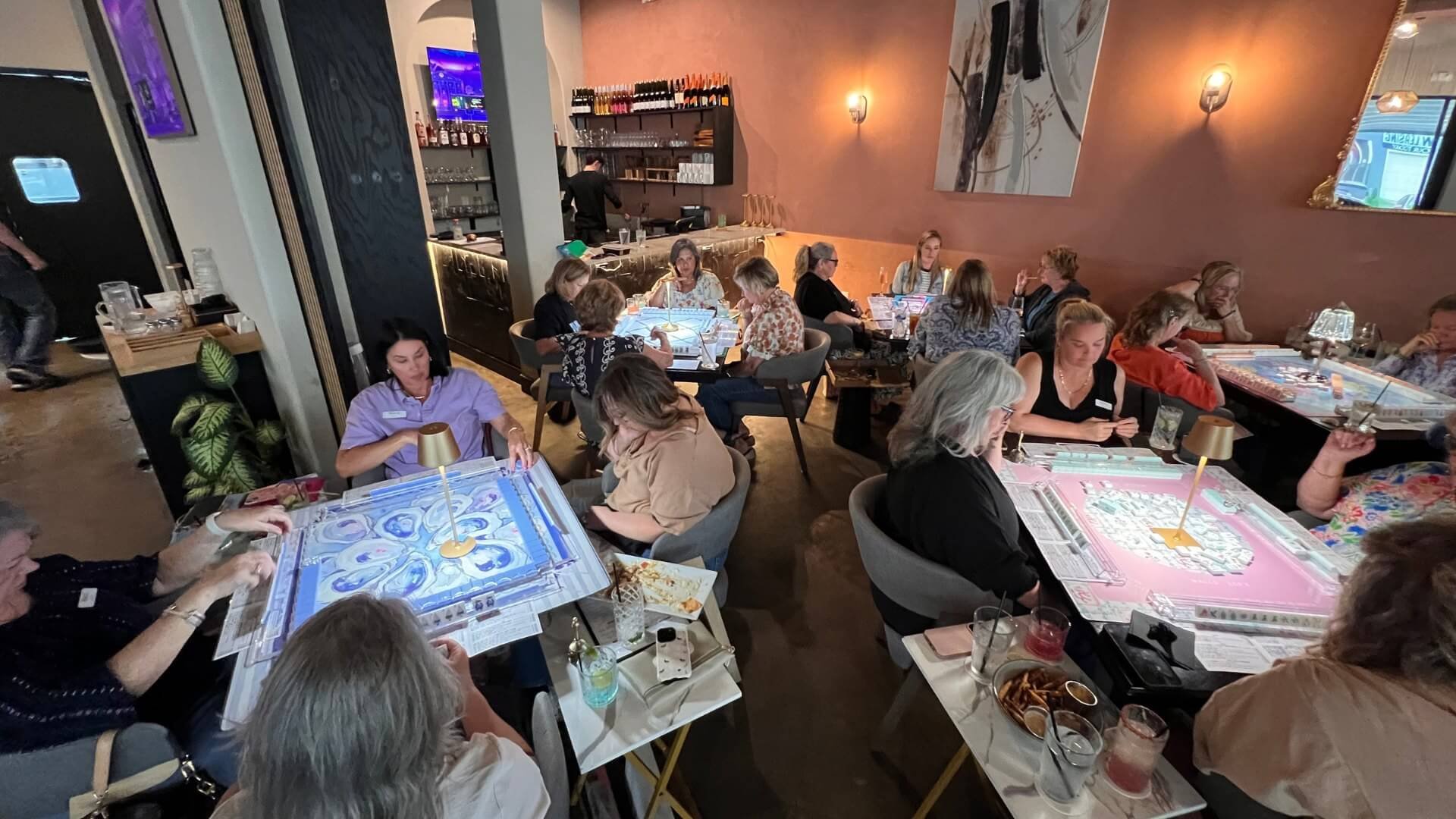 A group of people sitting at tables in a restaurant playing board games. The tables have colorful game boards and players are engaged in the game. The restaurant has warm lighting, modern decor, and a bar area in the background.