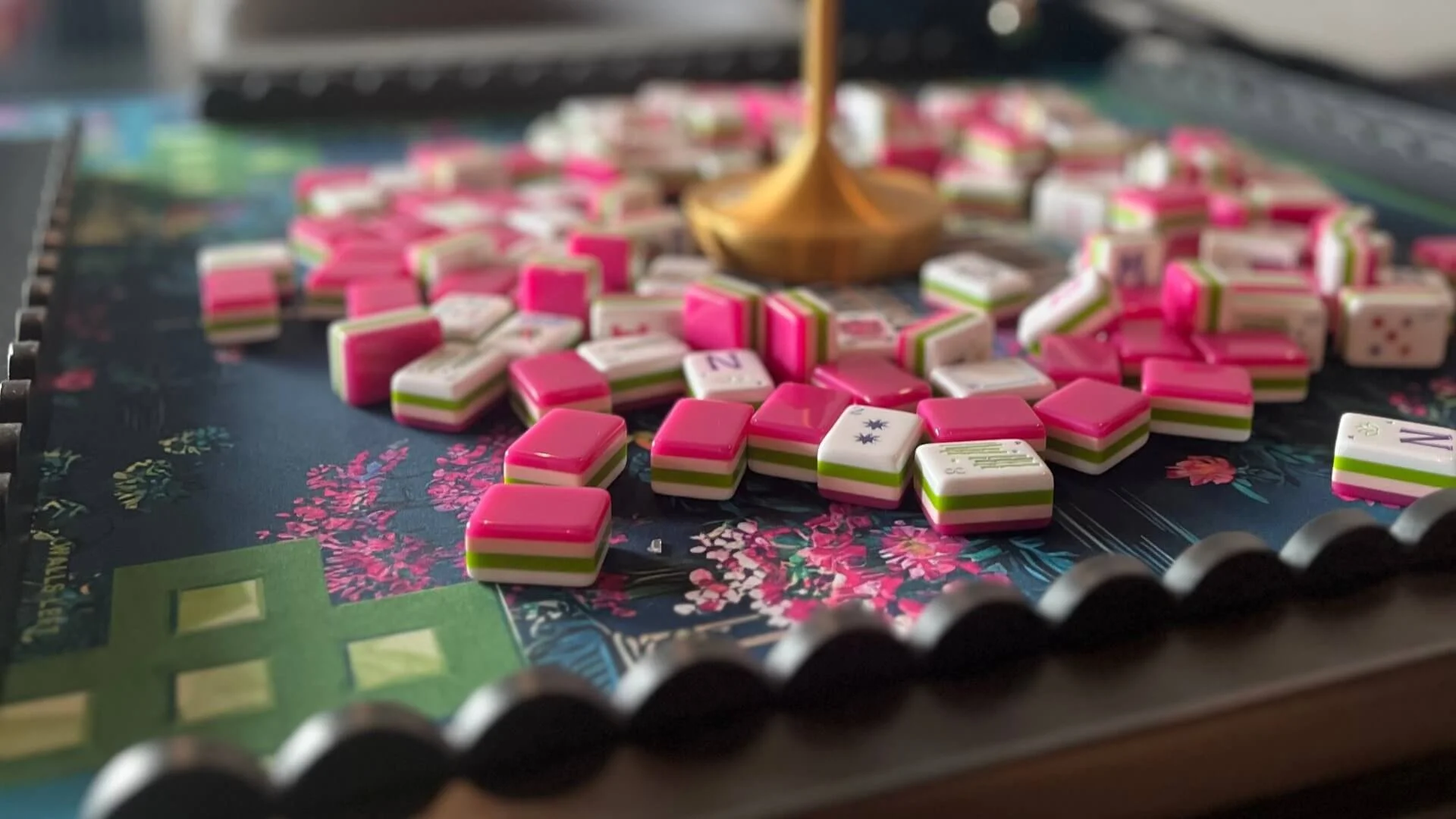 Close-up of a colorful board game with pink, white, and green tiles scattered on the game board, a stack of tiles near the edge, and a gold-colored game piece in the center.