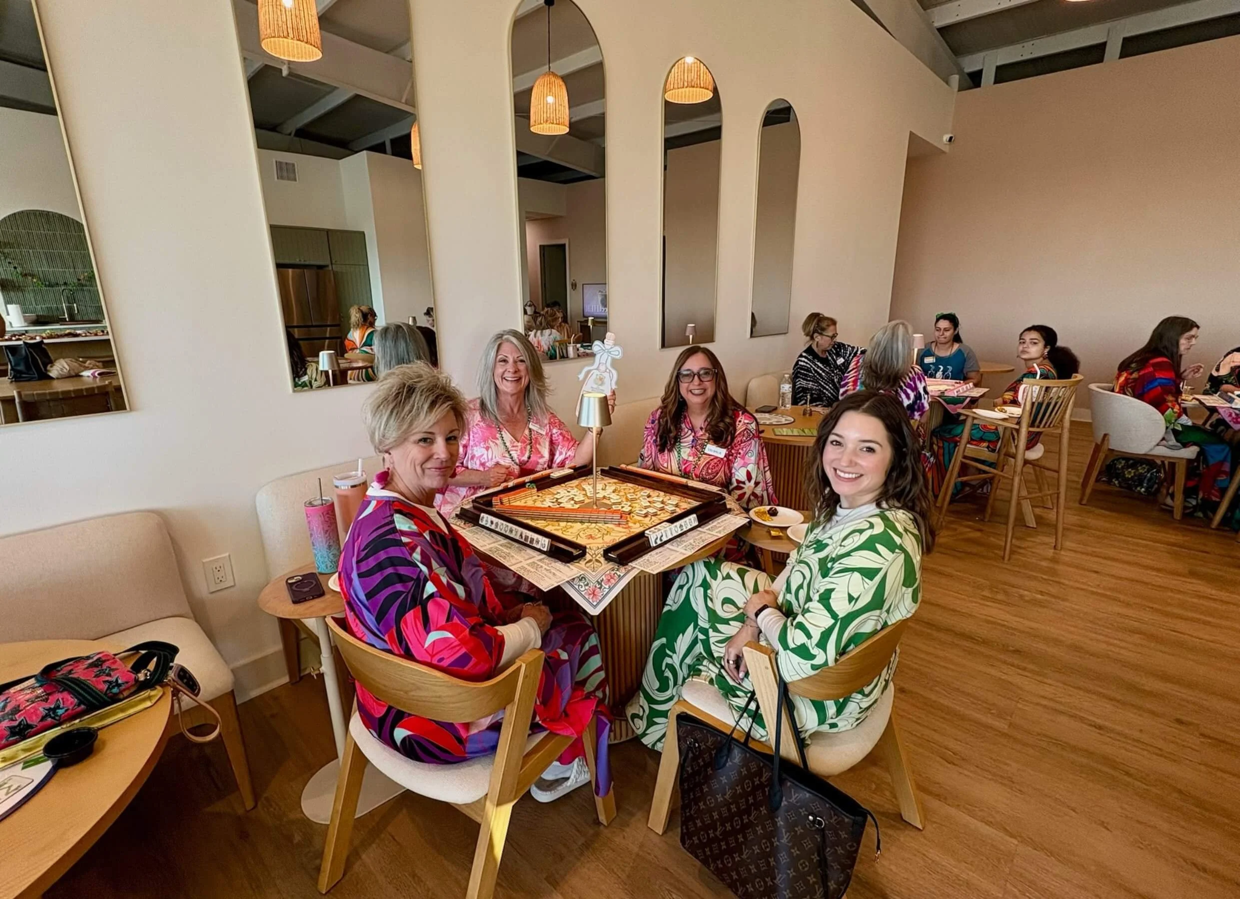Group of women enjoying a social gathering in a bright, modern restaurant, sitting around a table with a pizza and drinks, smiling at the camera.