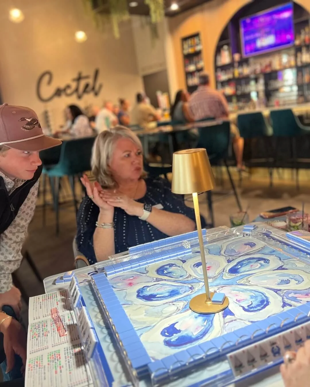 A woman with gray hair in a navy blue dress and a smartwatch on her wrist is sitting at a table, making a playful face with her lips puckered and hands near her face. The table features a decorative blue and white mahjong board