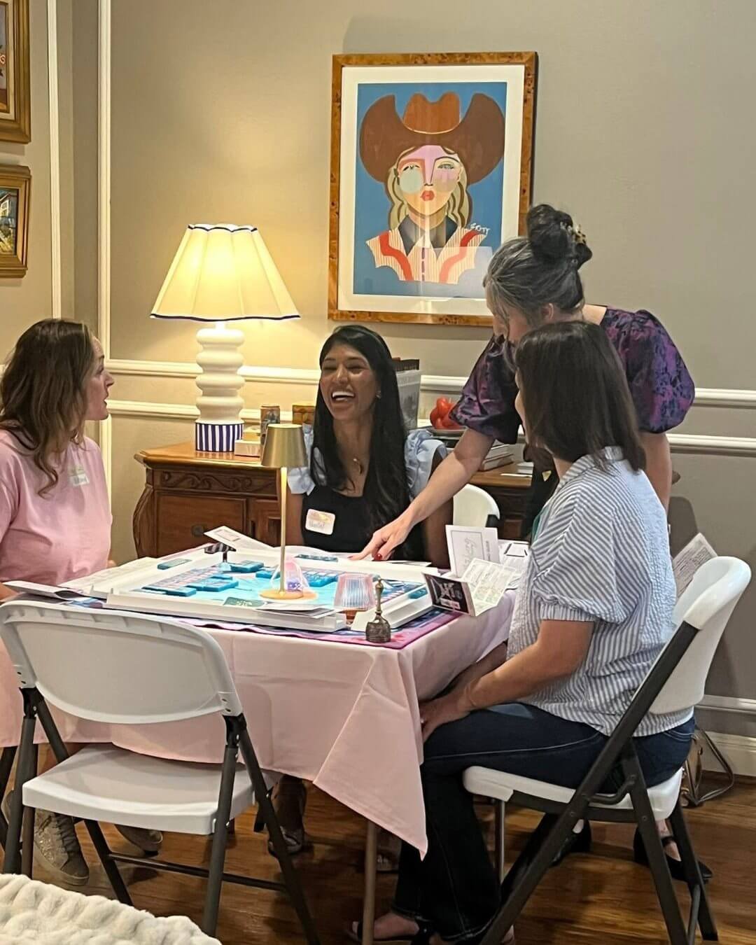 Four women are sitting around a table playing a board game in a cozy room with warm lighting, a lamp, and colorful artwork on the wall.