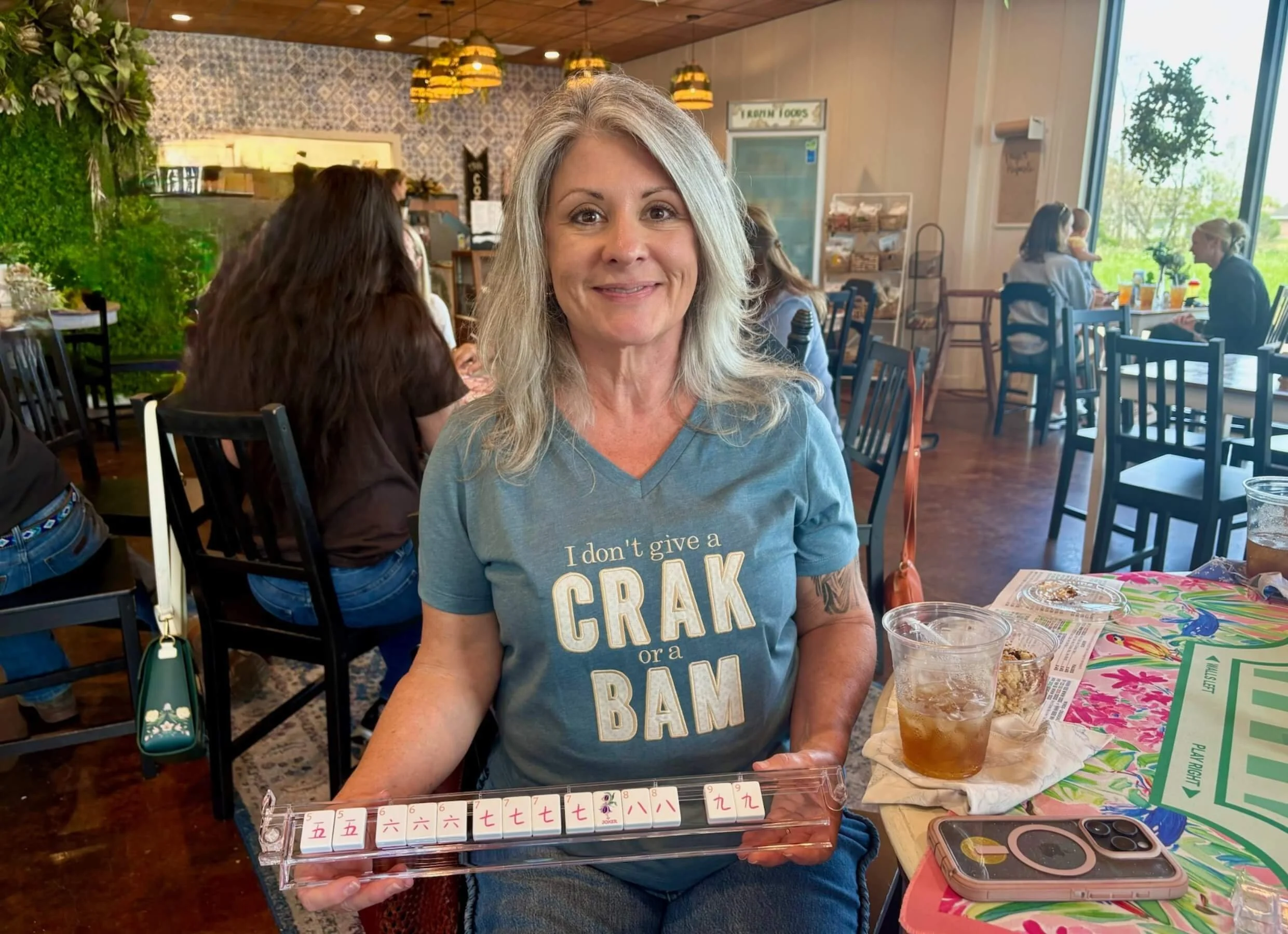 A woman with gray hair smiling at the camera in a restaurant. She is wearing a blue T-shirt with text and holding a Mahjong tile rack with numbered tiles in front of her. There are drinks, food, and colorful menus on the table. Other patrons are visible in the background, sitting at tables near large windows.