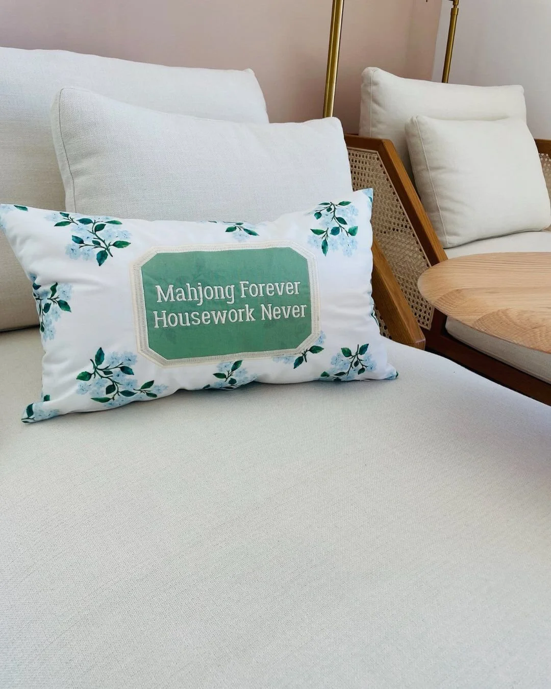 Decorative throw pillow with a humorous message, arranged on a white fabric sofa. The pillow has a floral border and reads 'Mahjong Forever Housework Never'.
