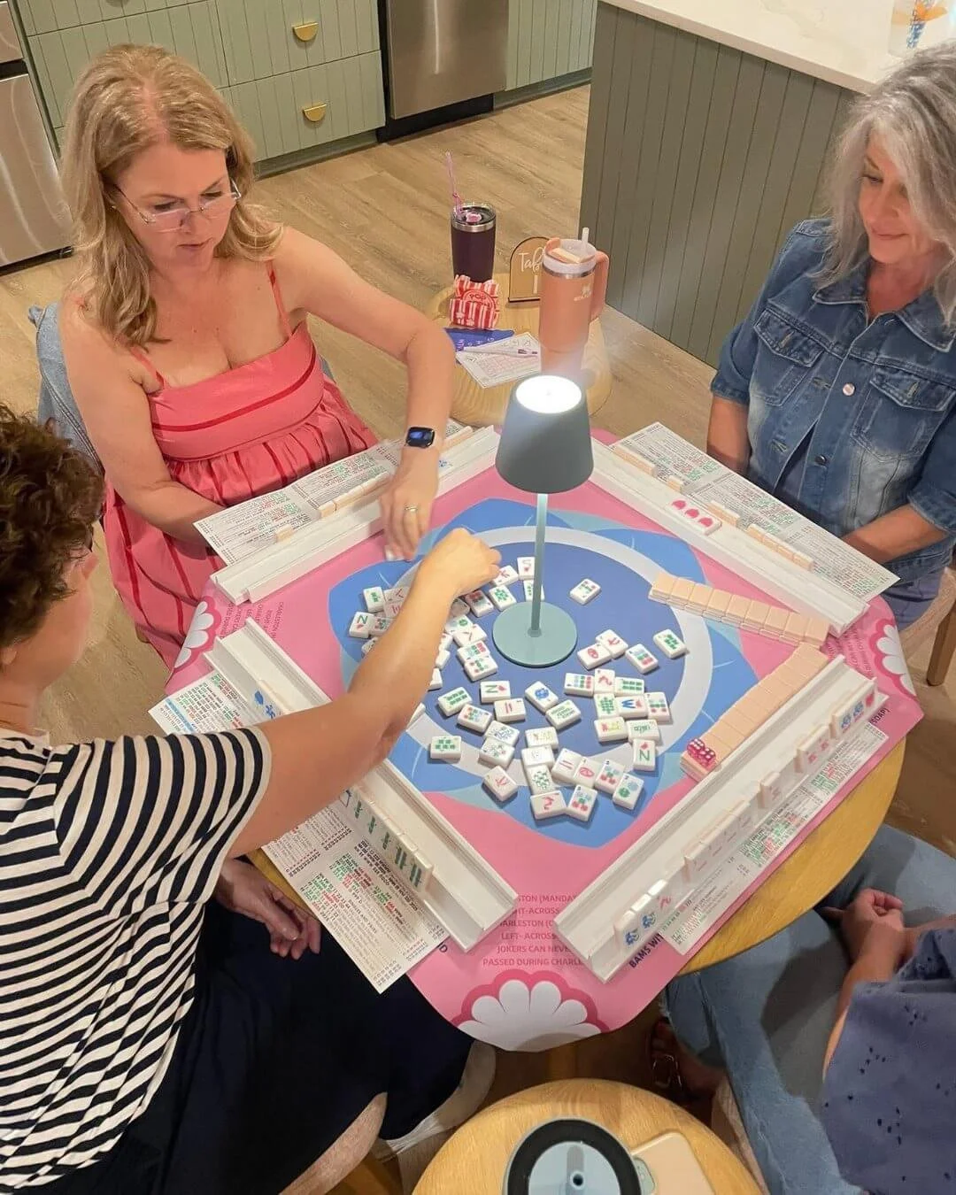Four women playing Mahjong at a round table in a cozy kitchen, using Mahjong tiles and a pink tablecloth with a lamp in the center.