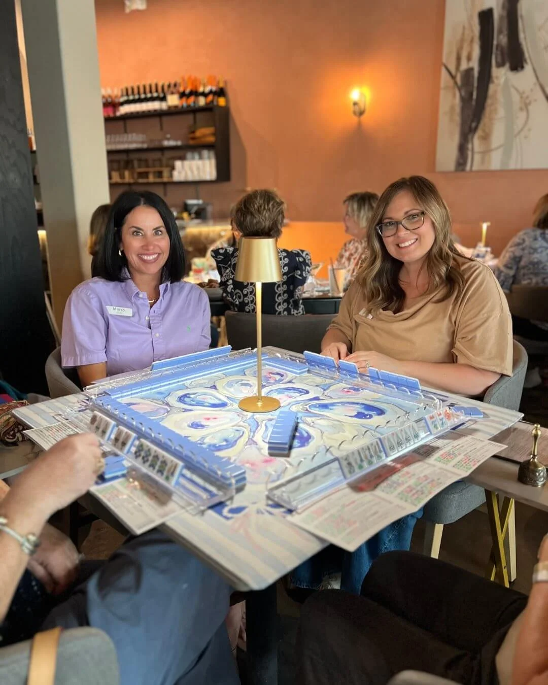 Two women playing a game of Mahjong at a restaurant table, smiling at the camera. The woman on the left wears a light purple shirt, and the woman on the right wears a beige top and glasses.