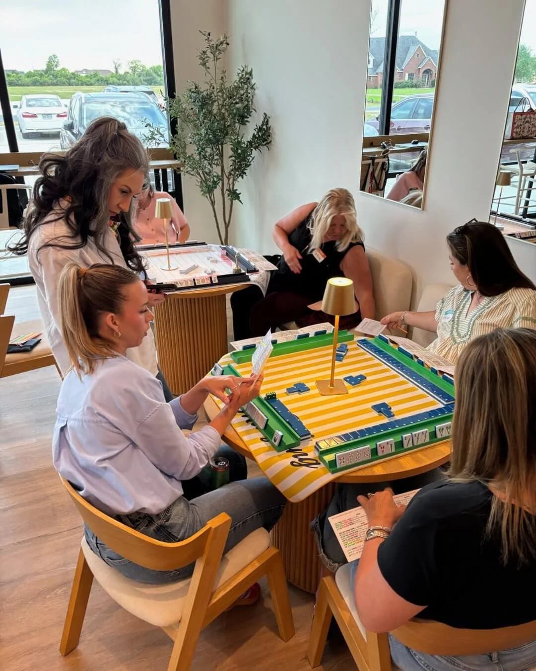 Women playing and viewing a game of Mahjong at a wooden table in a bright restaurant or cafe with large windows and parked cars outside.