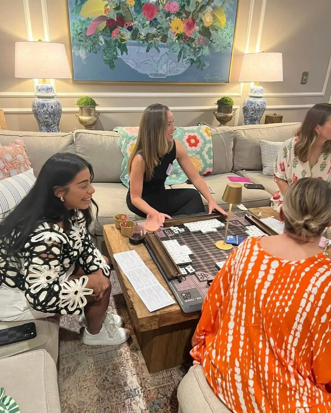Group of women learning to play mahjong at a cozy living room with artwork of flowers, table lamps, and decorative pillows.