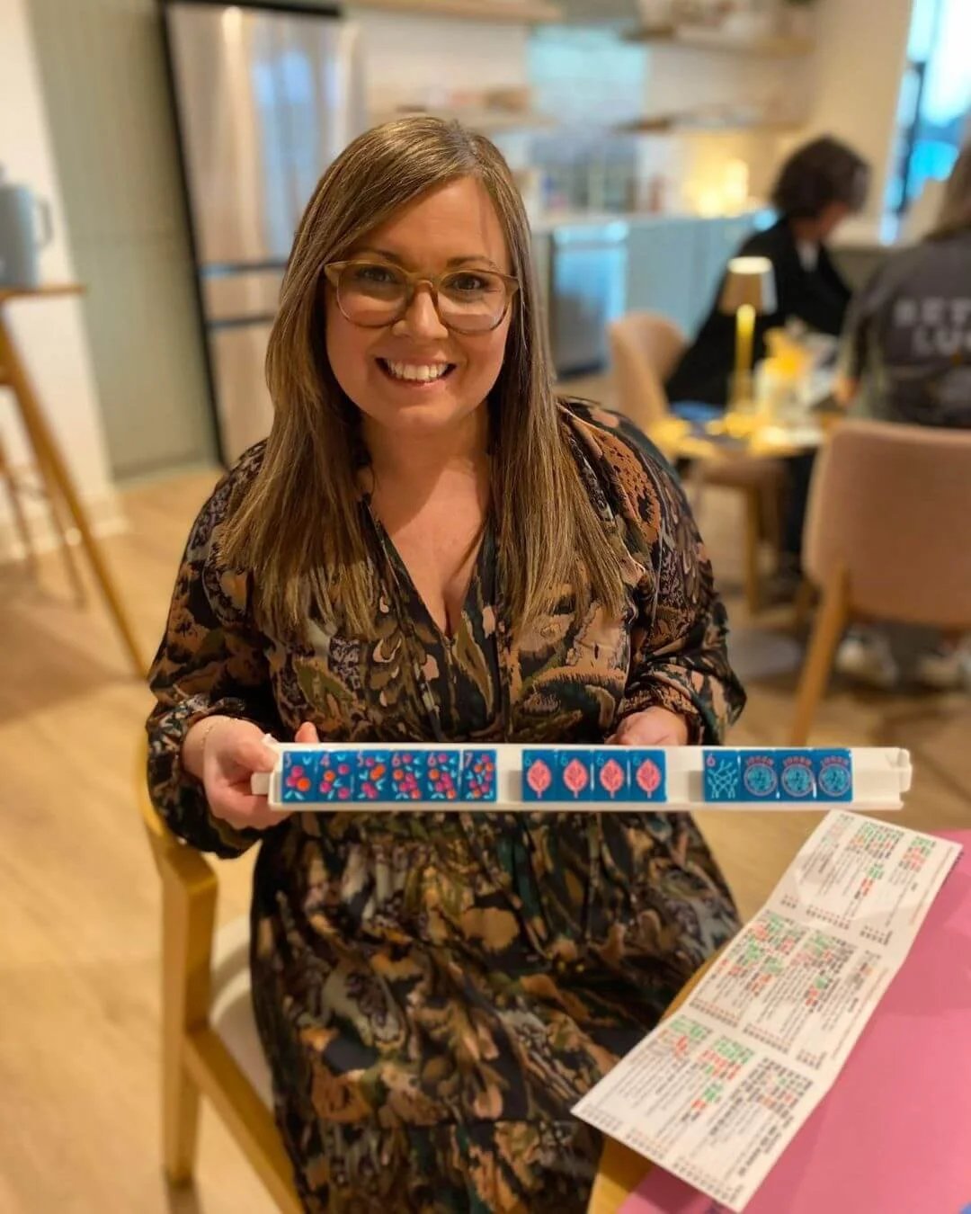 A woman with glasses and long brown hair smiling and holding a mahjong tile rack, sitting at a table in a restaurant.