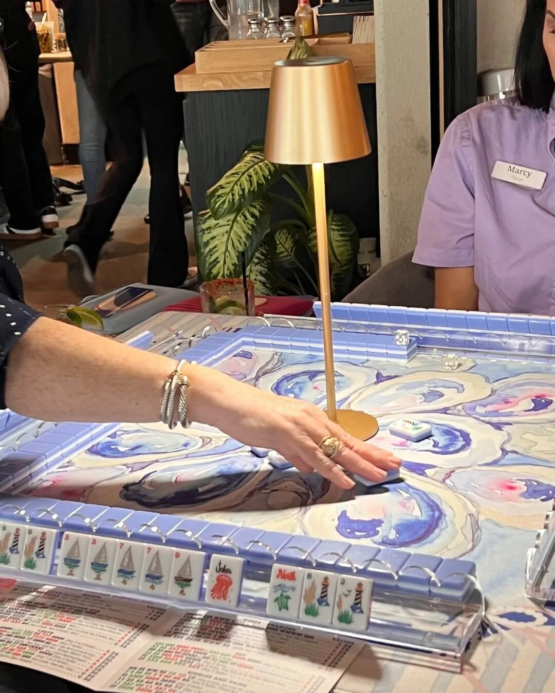People playing a game of Mahjong at a table with a decorative cloth, a lamp, and a potted plant in the background.