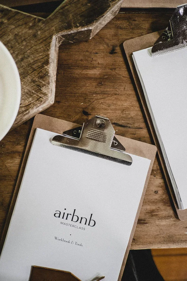 Caderno de anotações com o título 'airbnb MASTERCLASS Workbook & Tools' em uma mesa de madeira, com uma caneta e uma pequena maior.