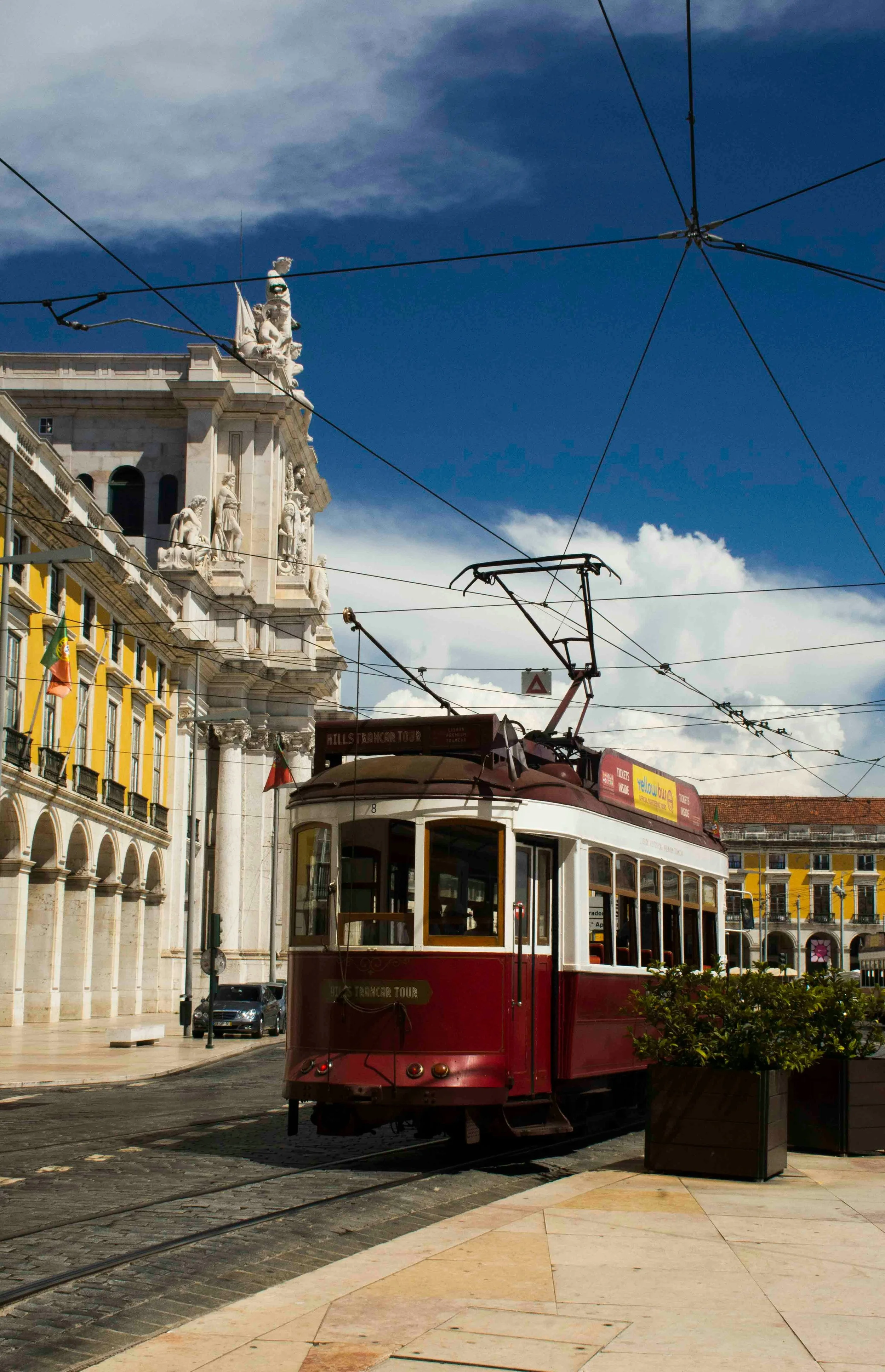 Elétrico vermelho em movimento na cidade, ao fundo edifícios históricos de arquitetura clássica, céu azul com algumas nuvens.