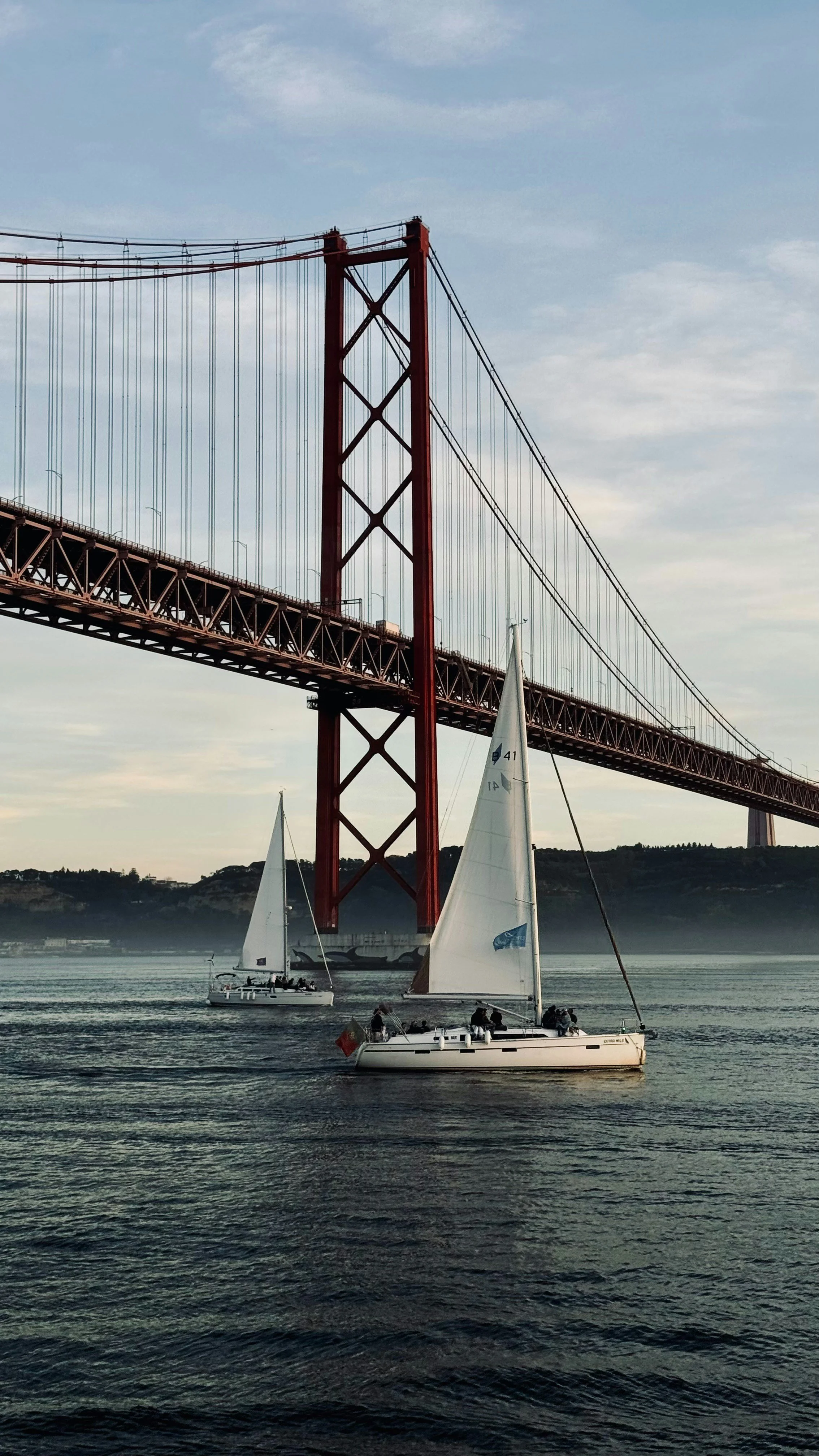 Ponte suspensa vermelha sobre um rio, com dois veleiros navegando na água.
