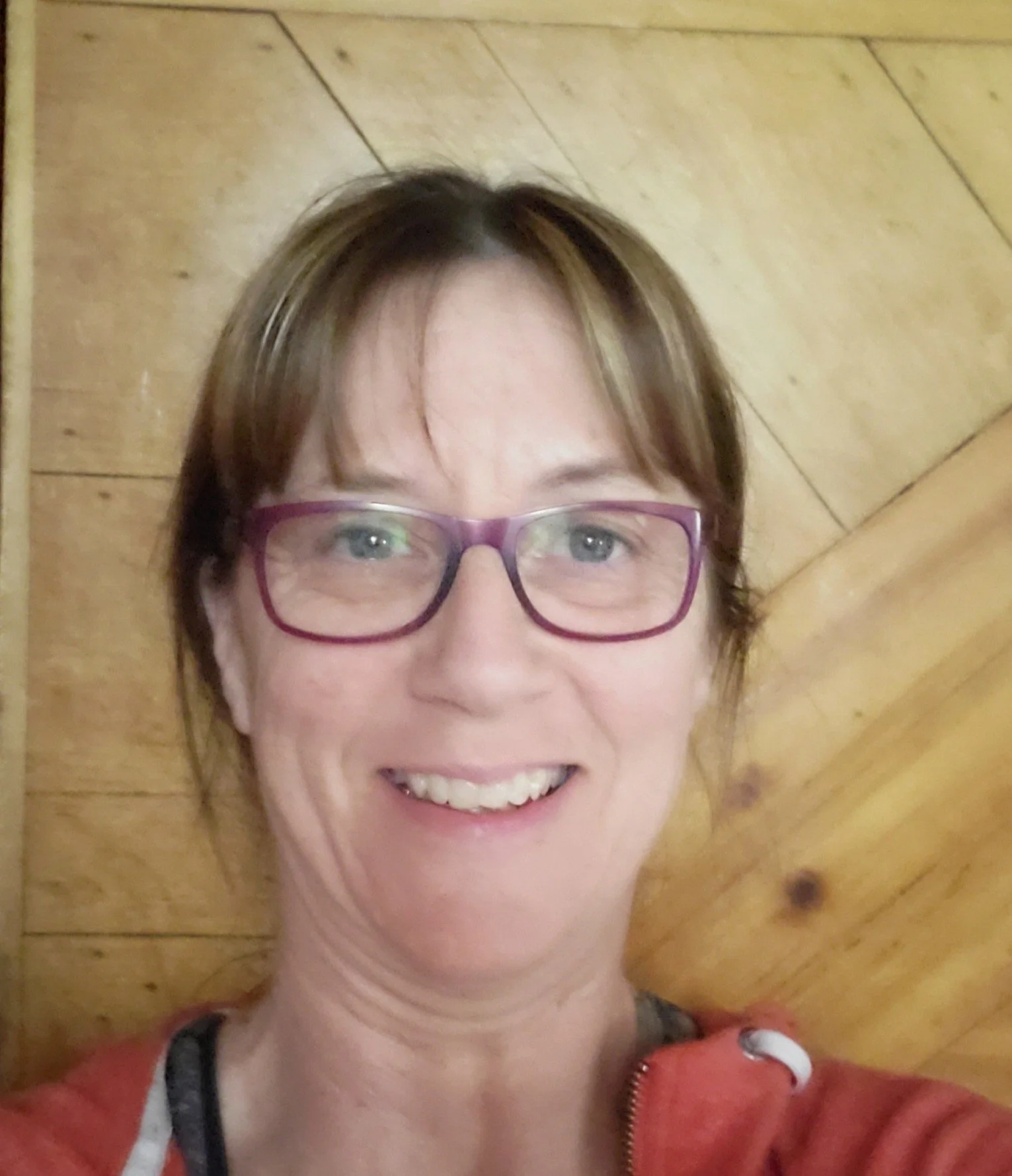 A woman with brown hair, purple glasses, smiling, in front of a wooden wall.