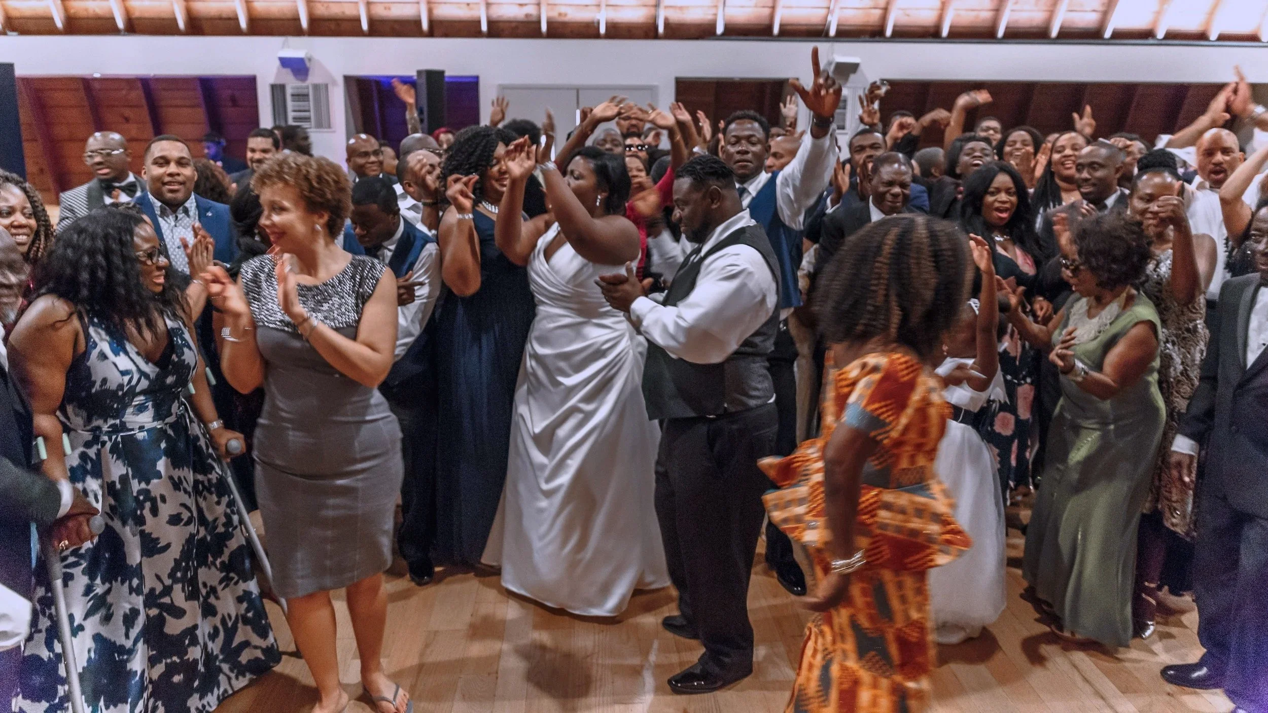 Group of people dancing, smiling, and celebrating in a room with wooden ceiling, dressed in formal and traditional attire.