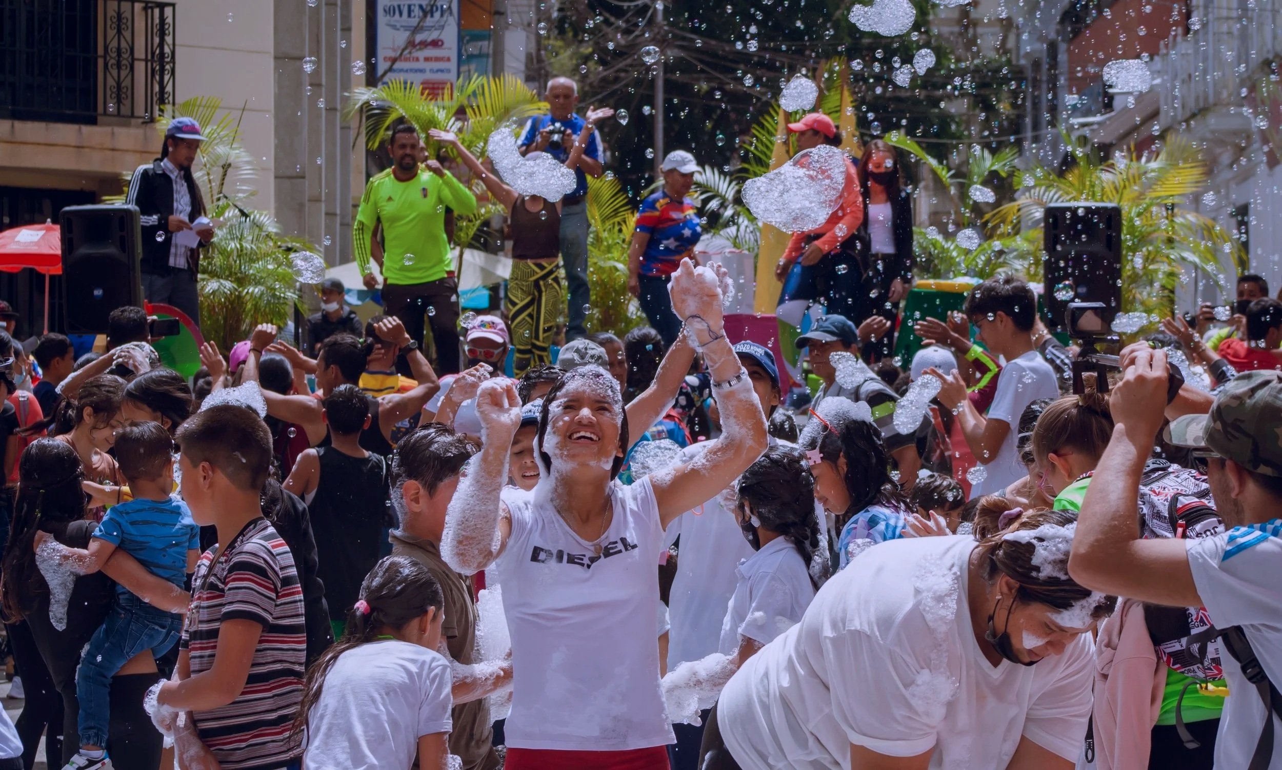 Crowd of people participating in a foam party on a city street, with some dancing and others taking photos, while foam bubbles fill the air and sunlight shines through.