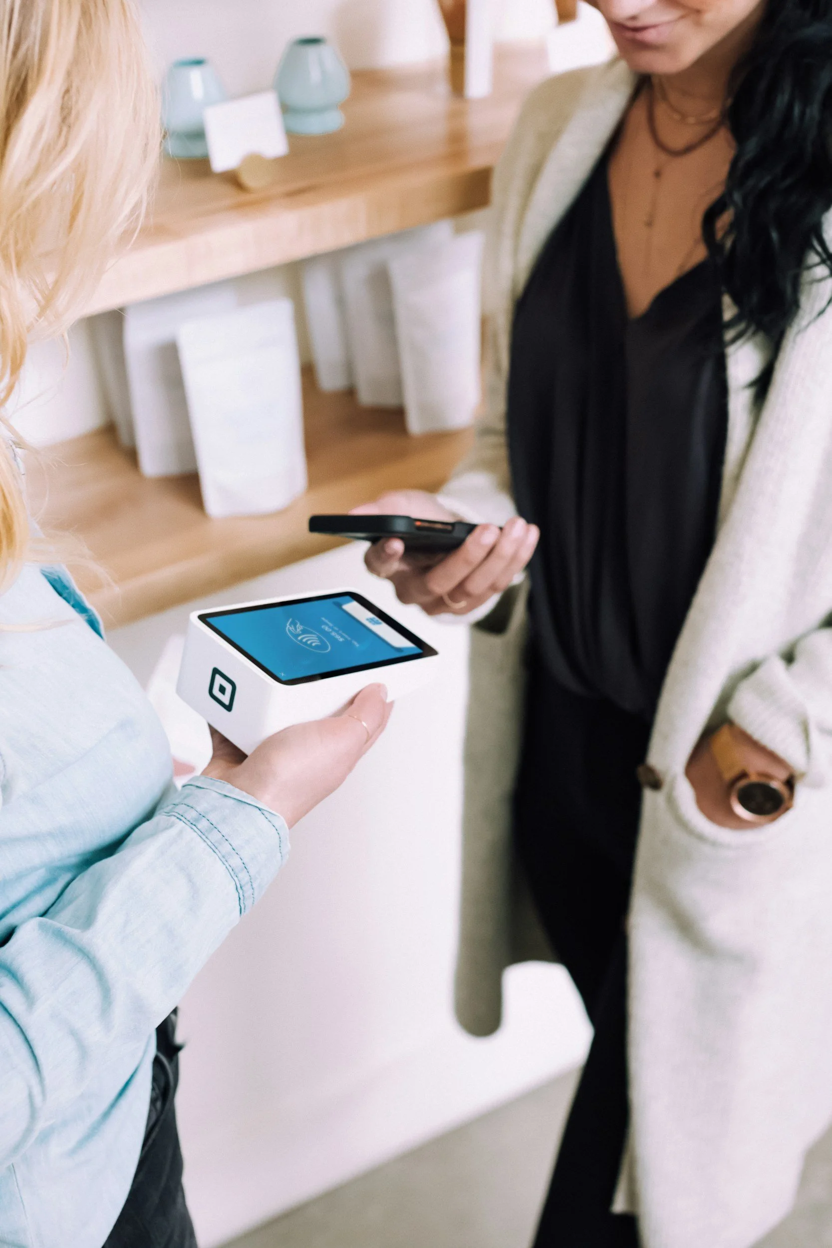 Two women at a checkout counter, one making a contactless payment with a smartphone, while the other holds a point-of-sale terminal.