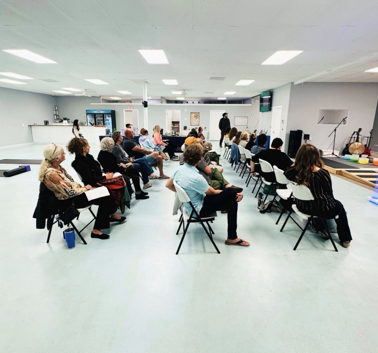 People seated in chairs facing a stage in a room, apparently attending a presentation or performance, with musical instruments on the right side of the room.
