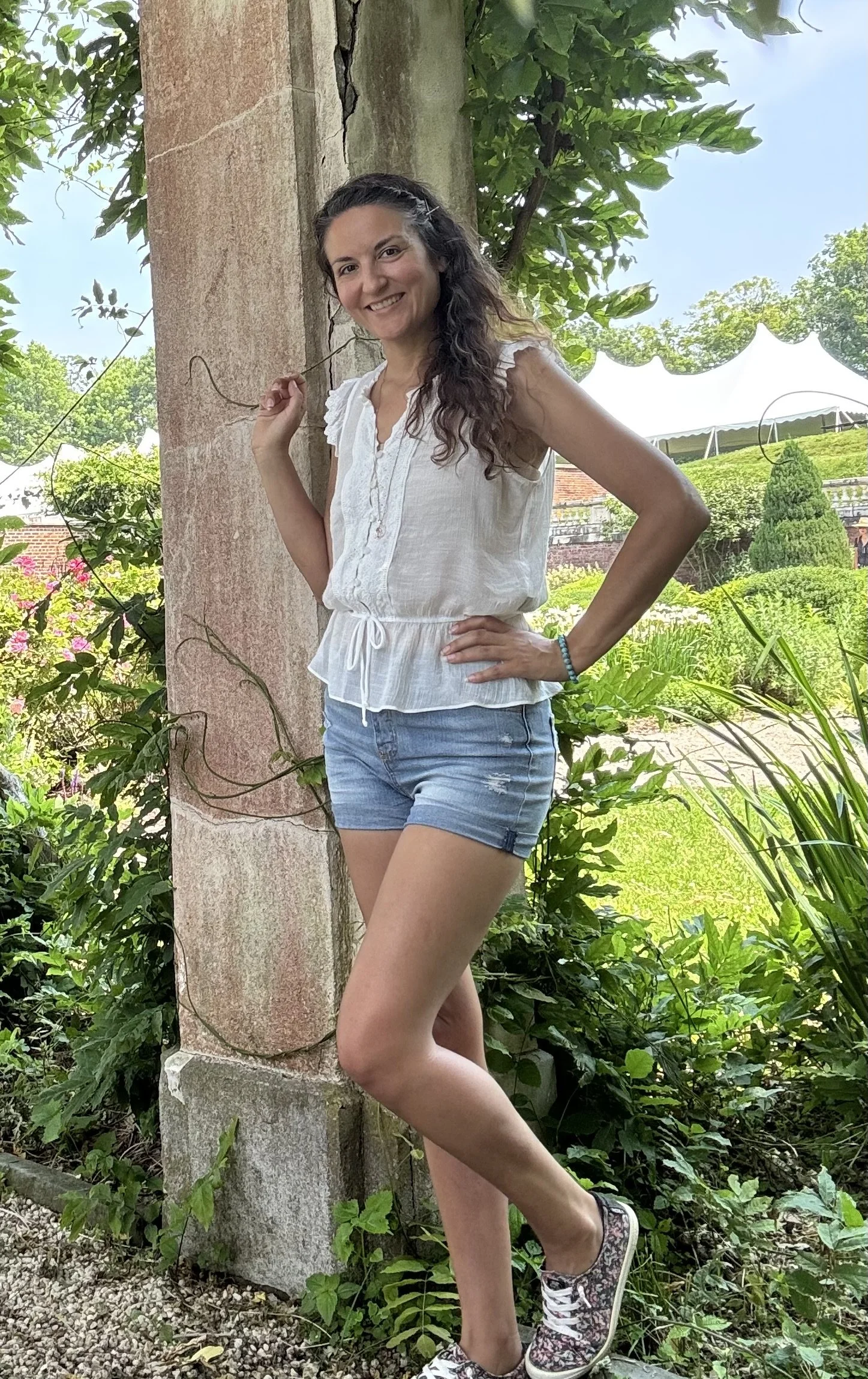 A woman smiling and posing outdoors, leaning against a tree with a lush green garden background, wearing a white sleeveless top, denim shorts, and sneakers.