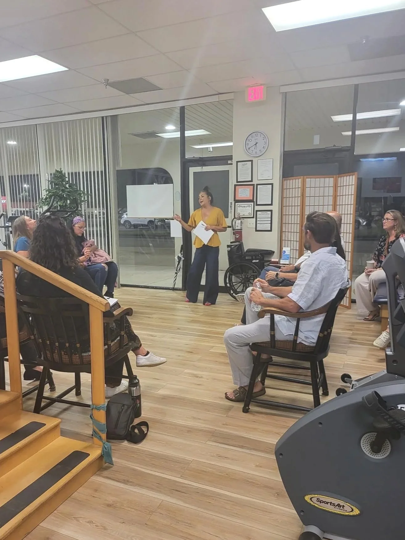 A group of people sitting in chairs facing a woman standing and speaking in a room with wooden floors, glass walls, and a sign reading 'EXIT'.