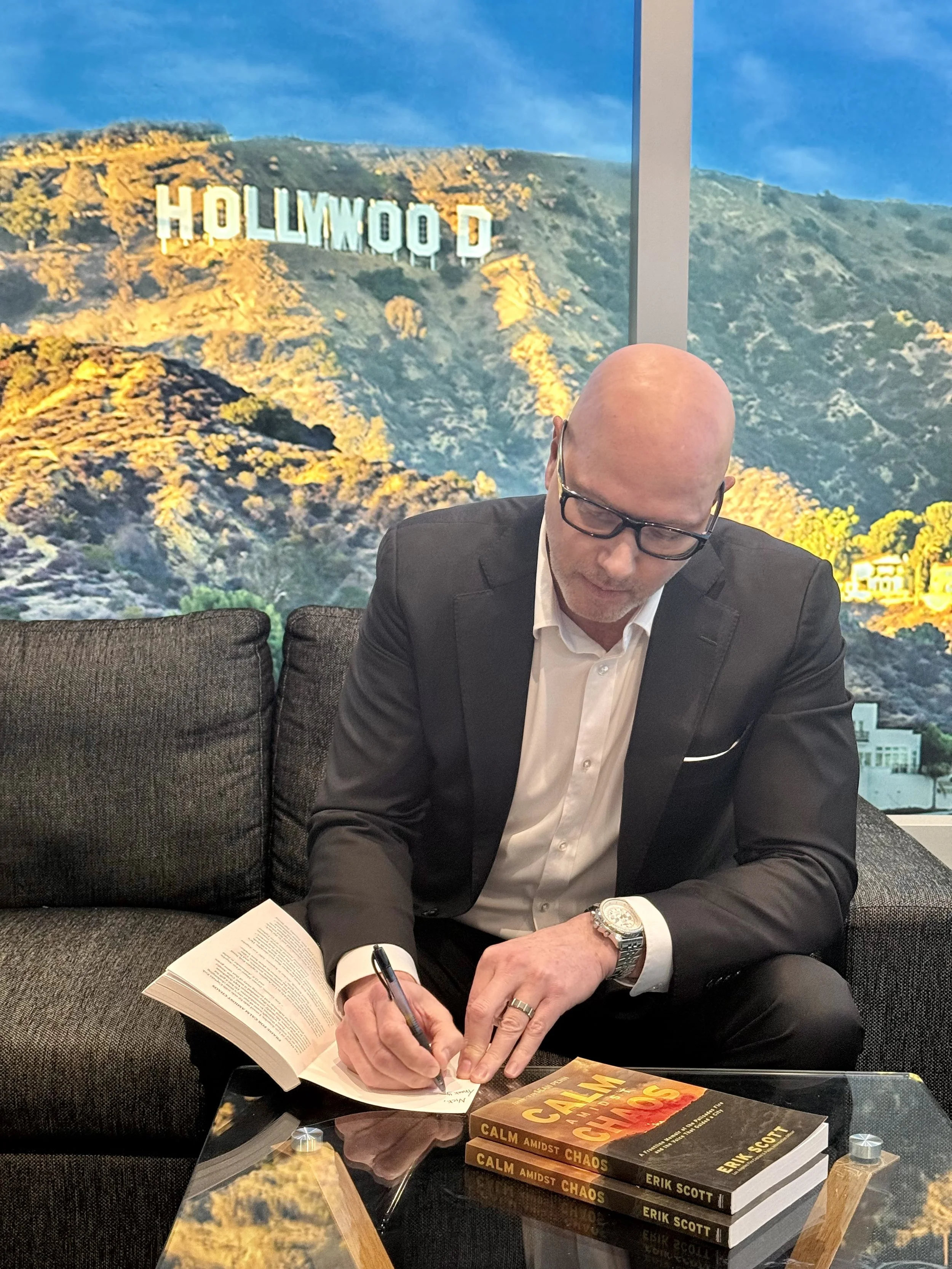 A man in a black suit with glasses, signing a book at a table with several copies of a book titled 'Calm Amidst Chaos' by Erik Scott. A large window behind him shows the Hollywood sign on a hill against a blue sky.