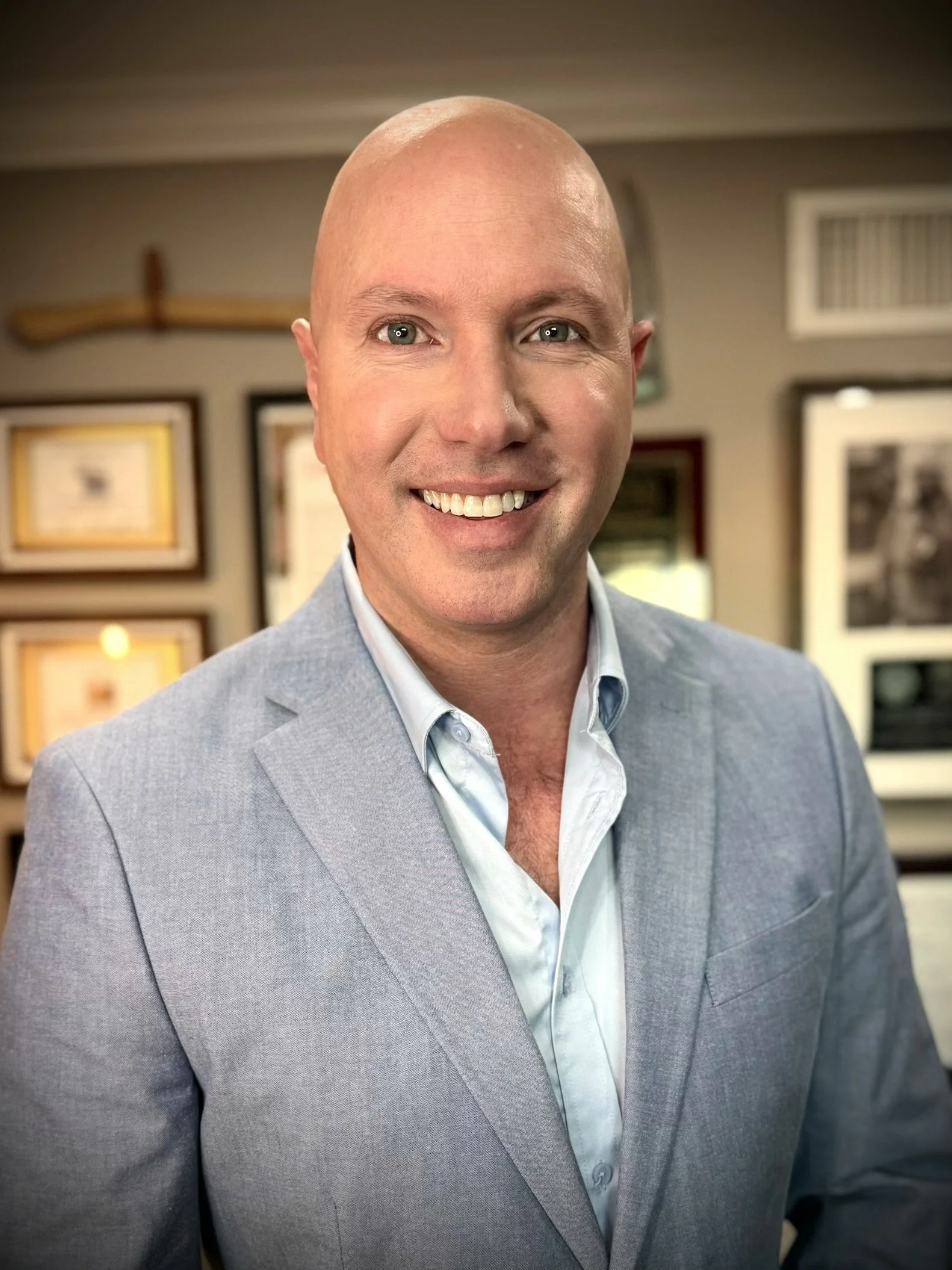 A smiling, bald man in a light gray suit and white shirt, standing inside a room with framed pictures on the wall behind him.