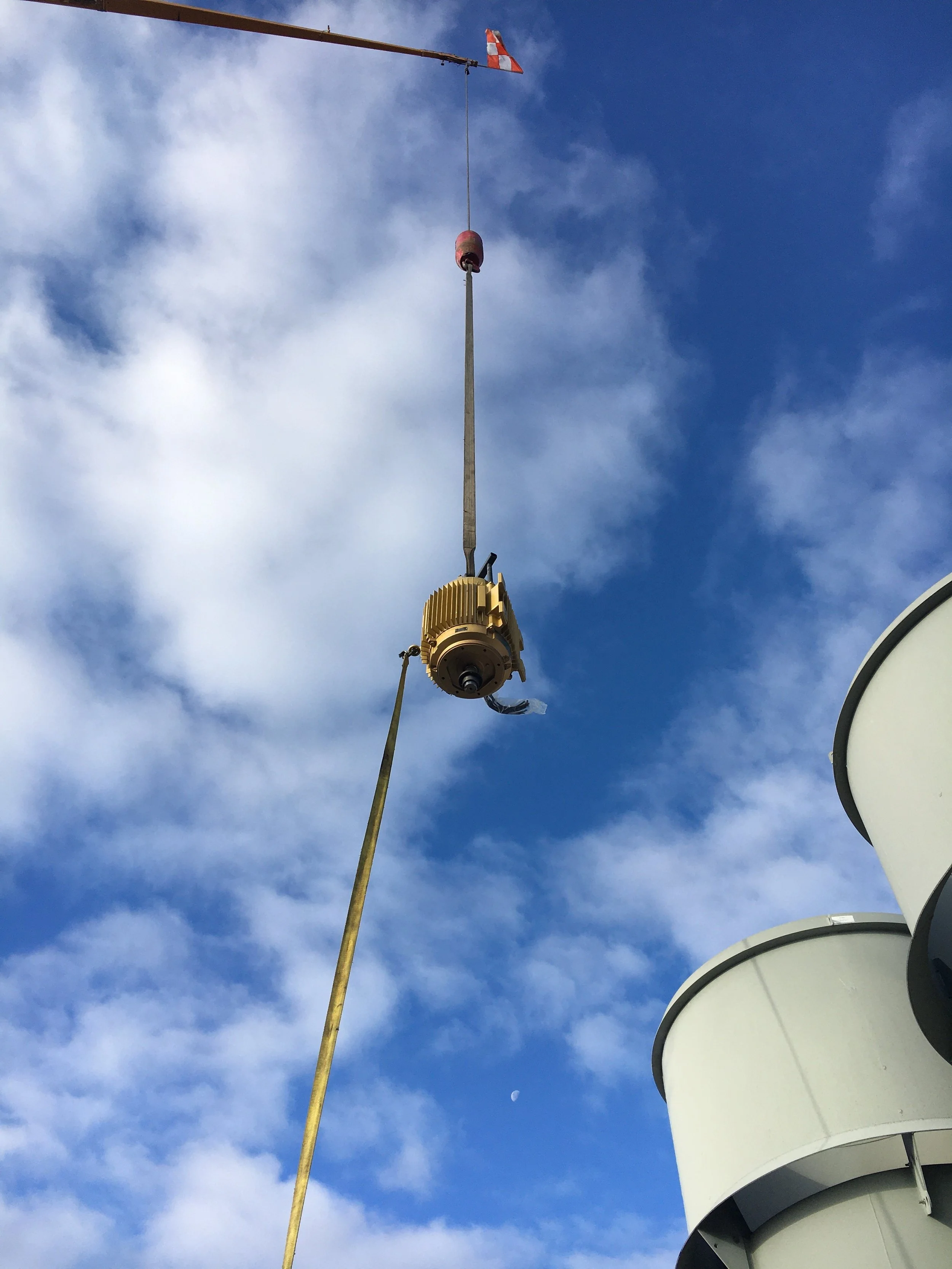 A construction crane lifting a large, yellow, cylindrical object late in the day with a bright blue sky with scattered clouds and a visible moon in the background.