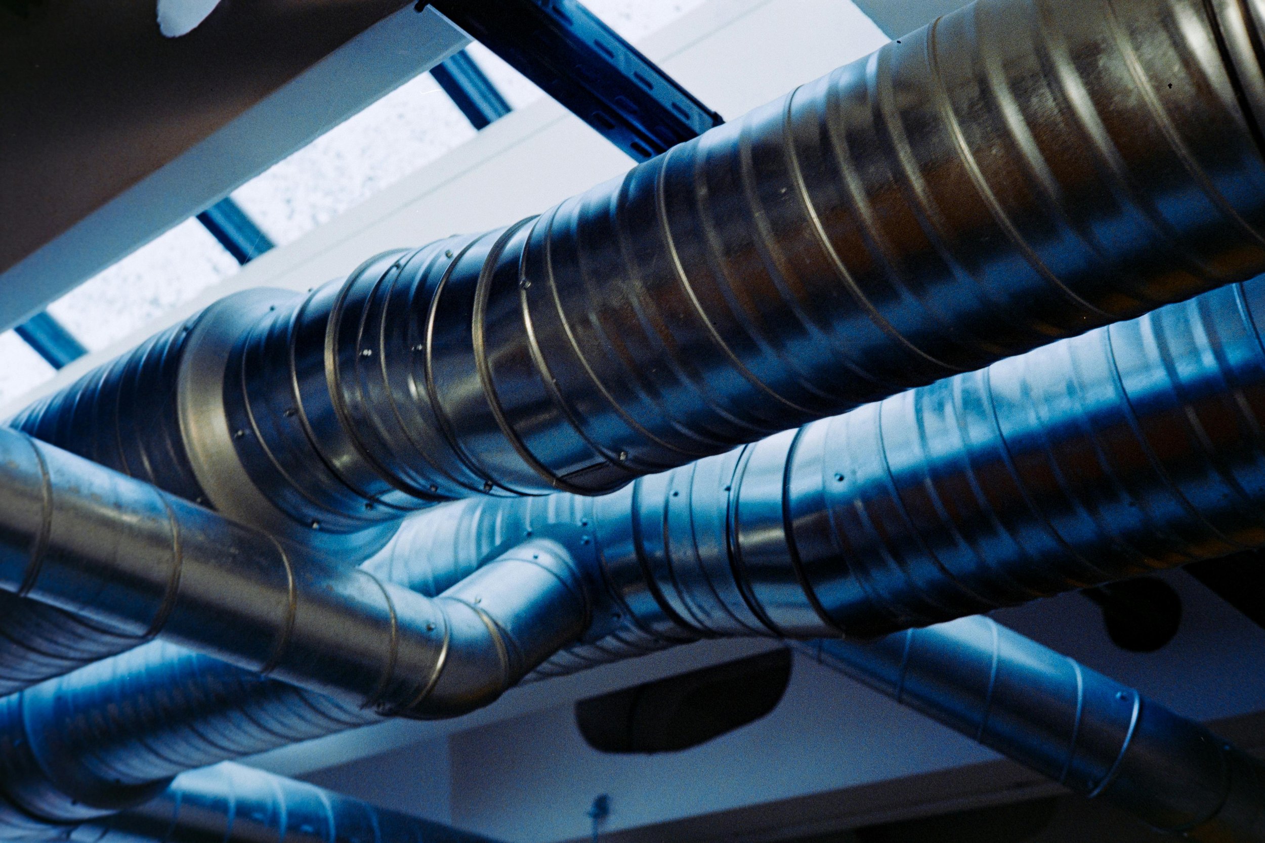 Close-up of metal ductwork pipes in a building ceiling, with some pipes appearing shiny and reflective, arranged below a skylight.