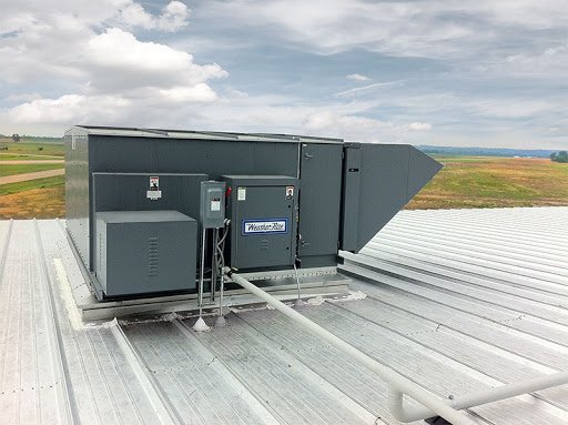 Industrial rooftop HVAC unit with a metal electrical box and ducts, set against a cloudy sky and open fields.