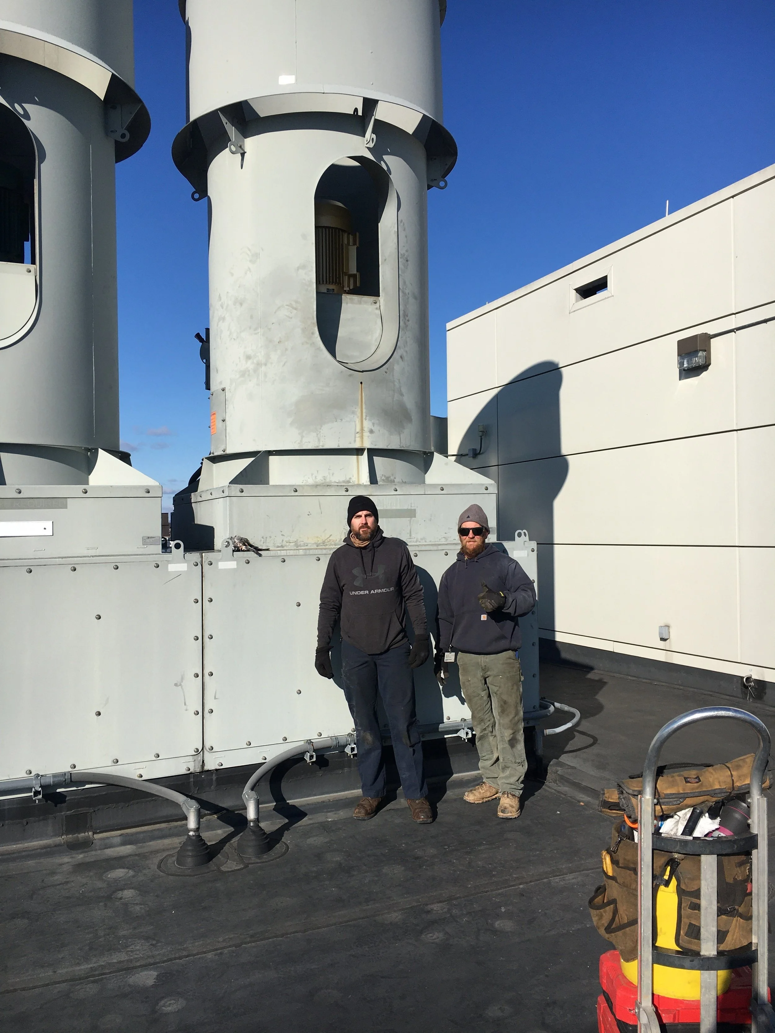 Two men standing on a rooftop with large industrial HVAC units and equipment around them, one giving a thumbs-up and both dressed in work clothes.
