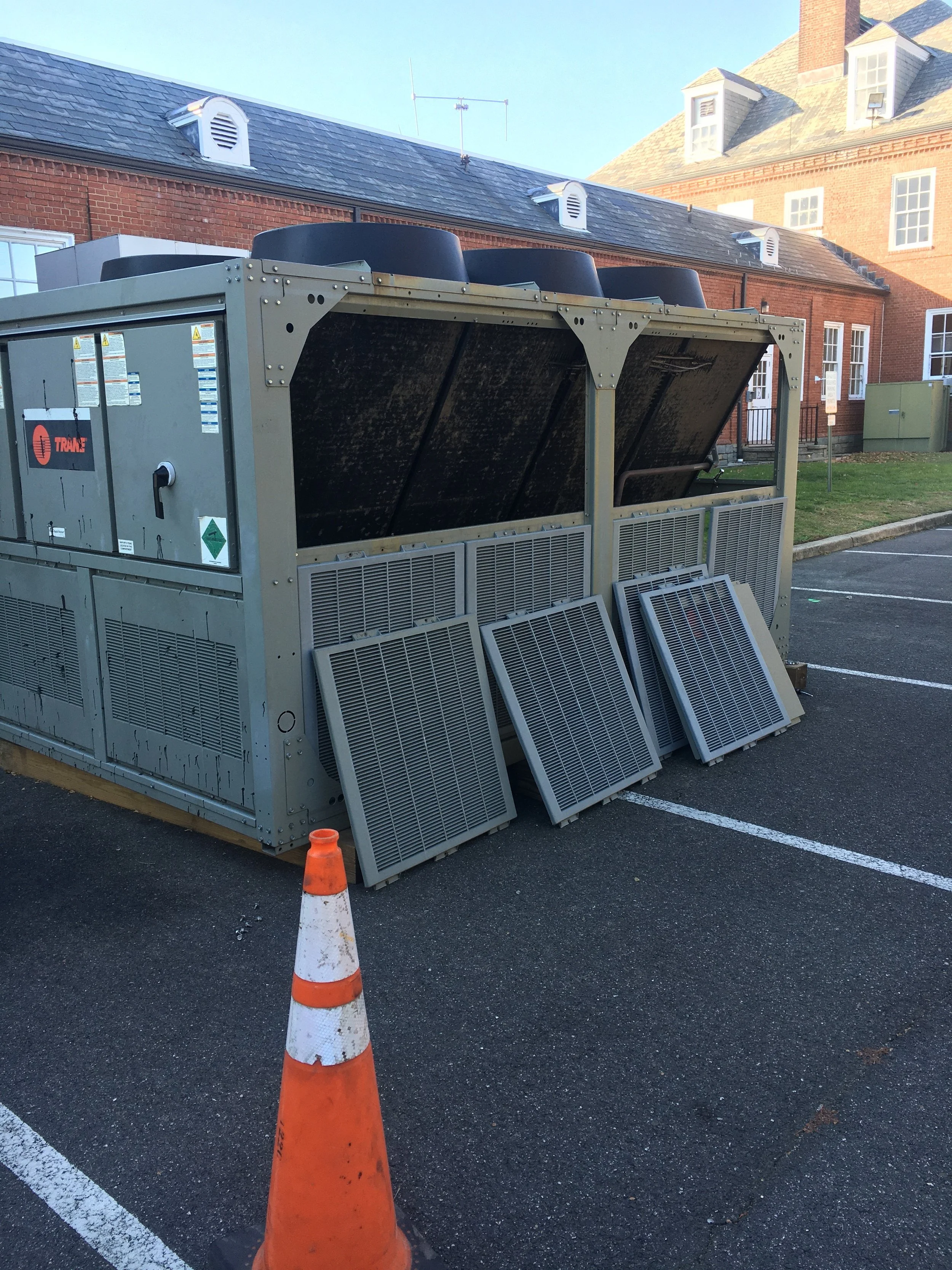 Large HVAC unit with several vent covers leaned against it, located in a parking lot near brick buildings, with an orange traffic cone in the foreground.